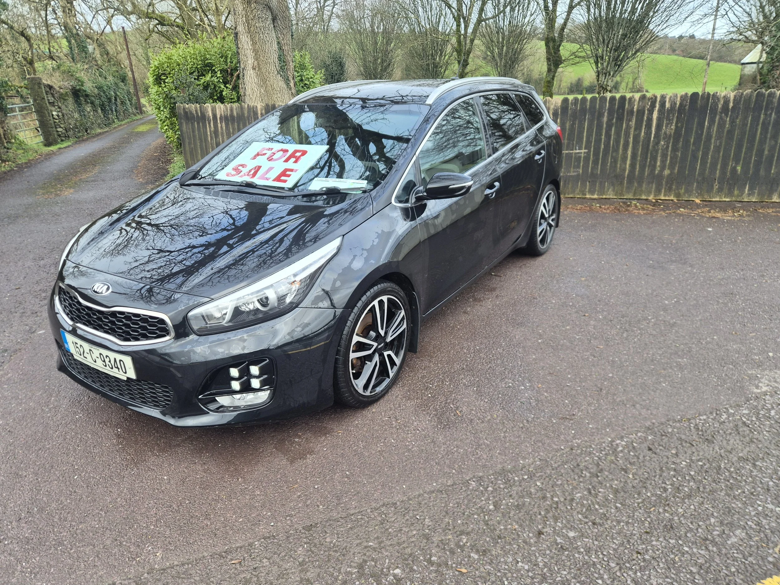 Black Kia sedan with a 'For Sale' sign in the windshield, parked on a gravel driveway surrounded by trees and a wooden fence, with a rural landscape in the background.
