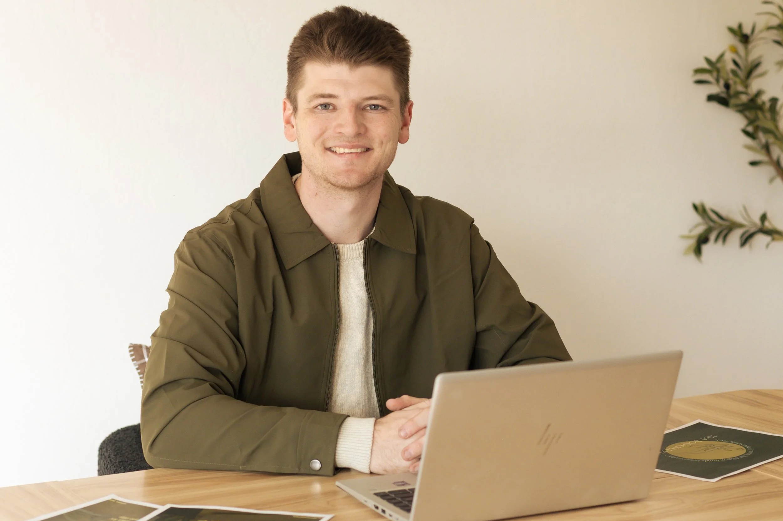 A young man with brown hair wearing a green jacket and beige sweater seated at a wooden table with a laptop and printed photos, smiling at the camera.