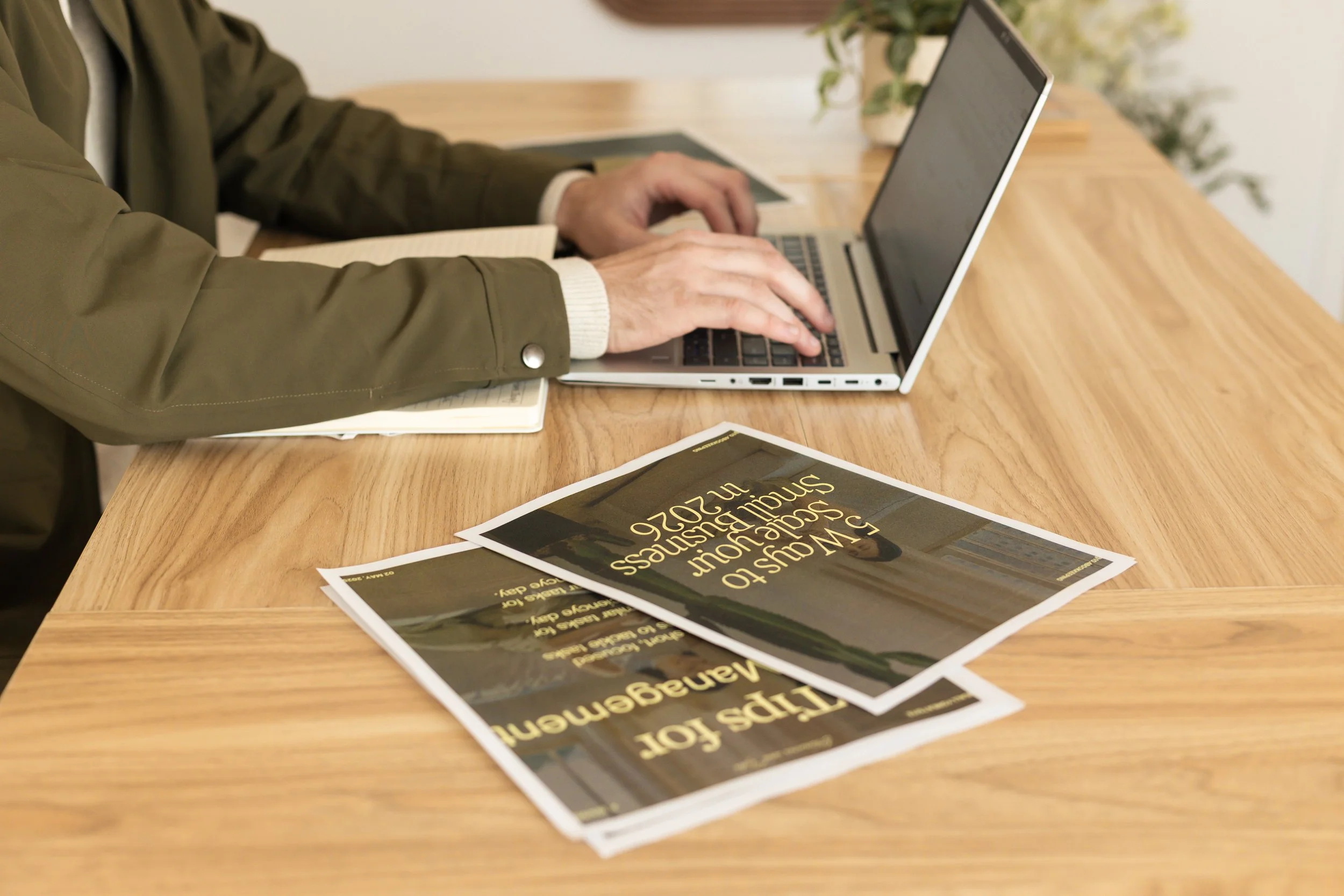 Person using a laptop at a wooden table with printed advertisements and a notebook around.