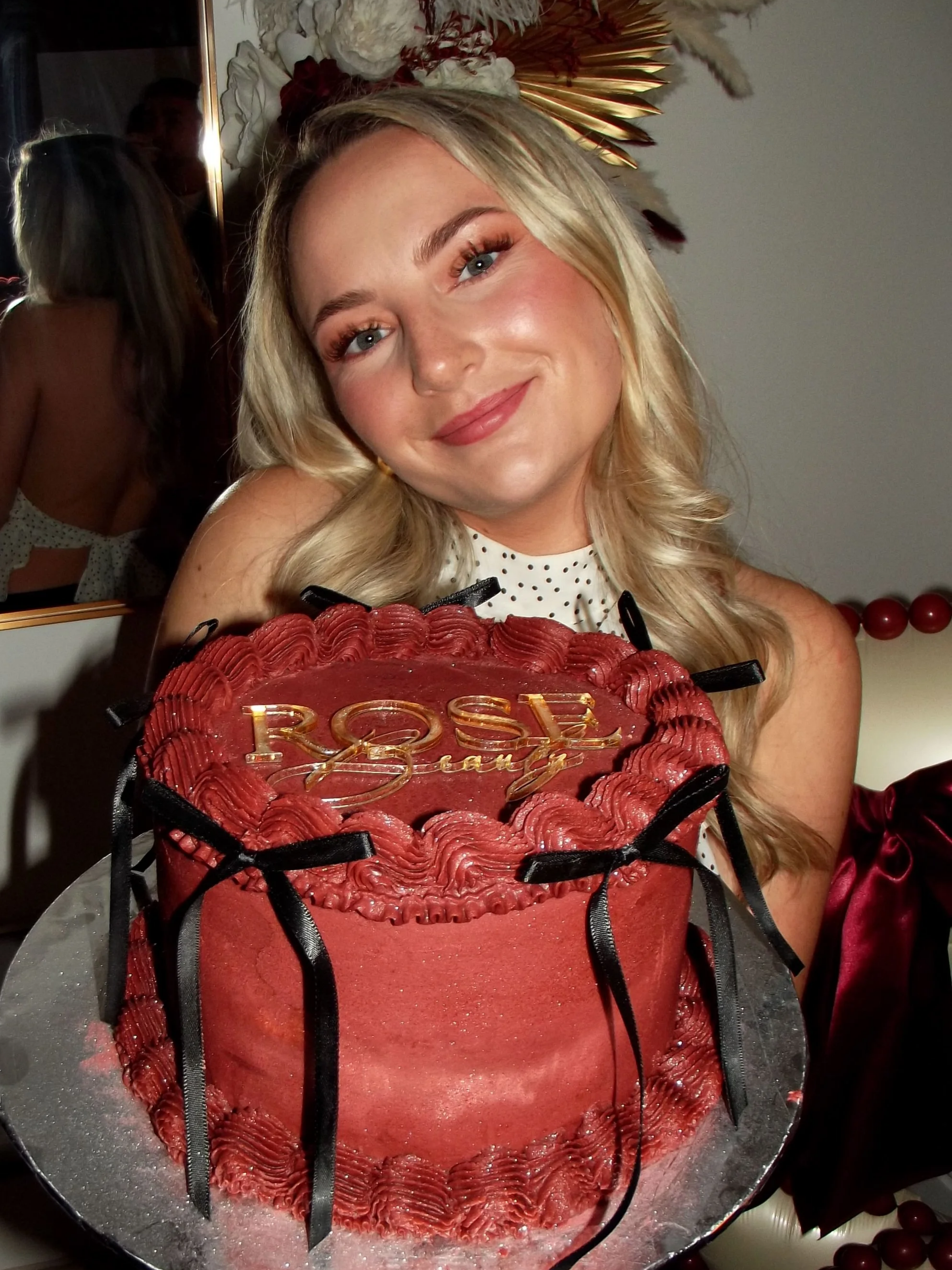 Blonde woman posing behind a large red birthday cake decorated with black ribbons and gold "Rosé" lettering.