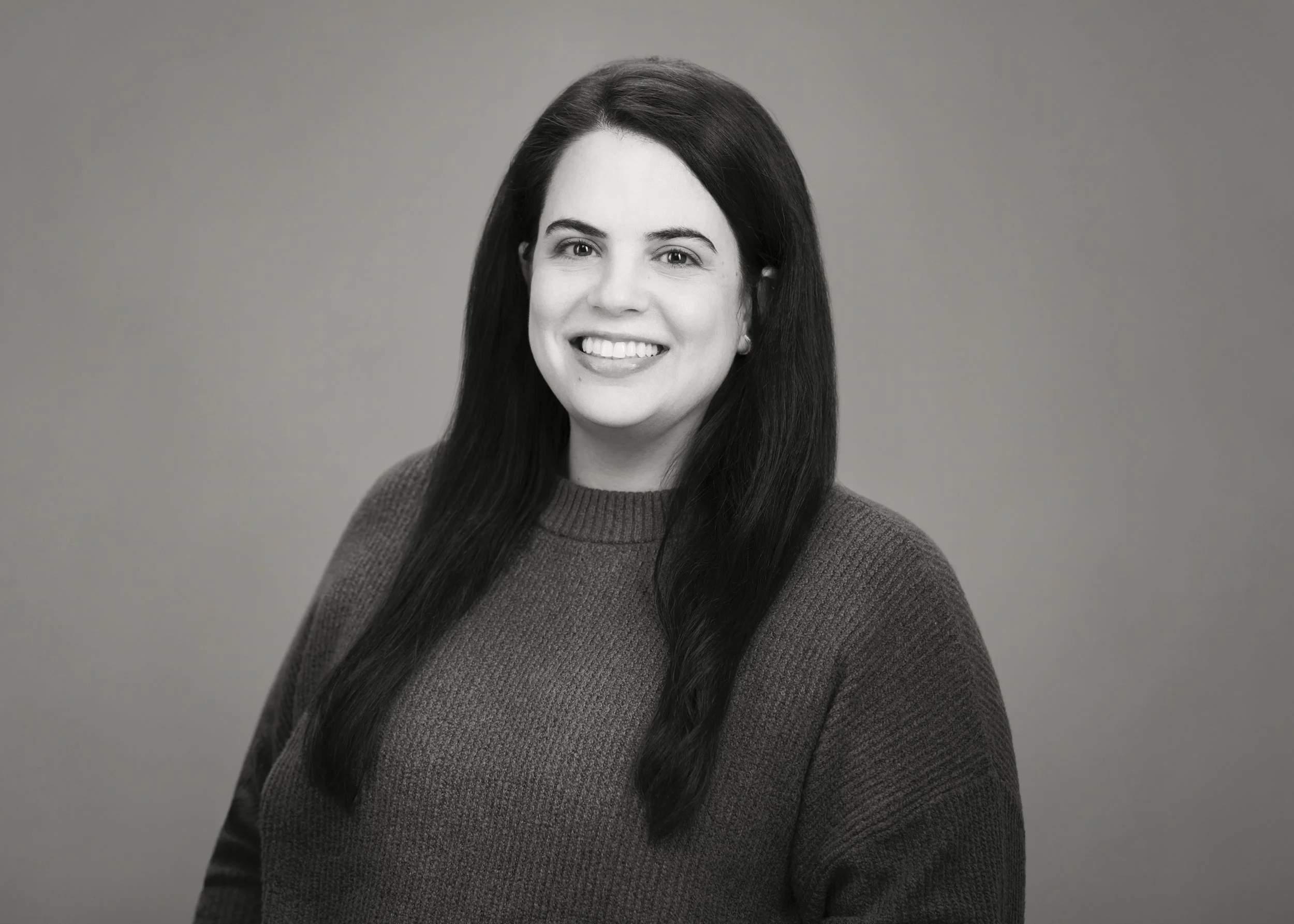 Black and white portrait of a woman with long dark hair, smiling, wearing a dark sweater, against a plain background.