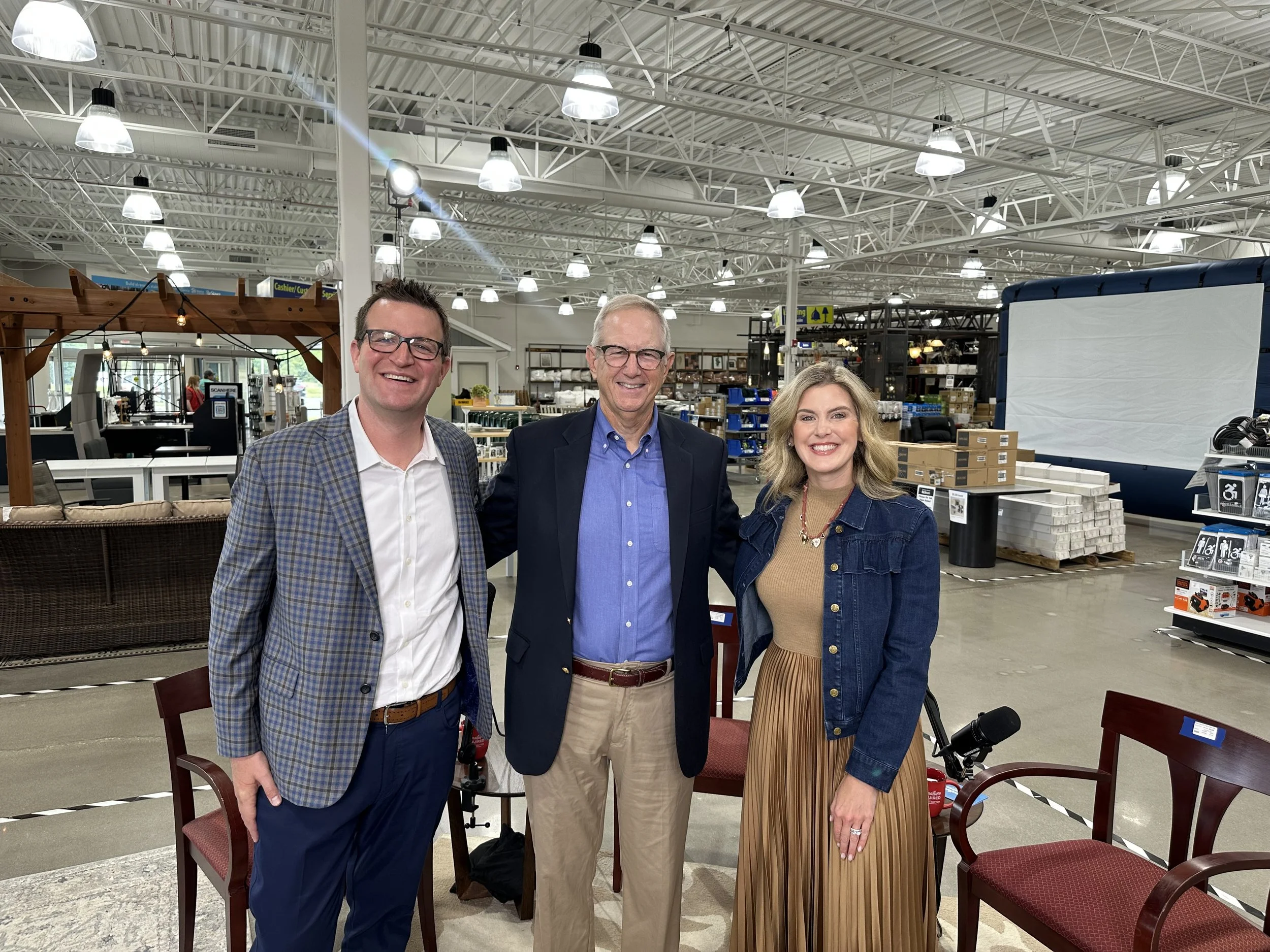 Three people standing in an indoor retail store, smiling at the camera. Behind them are tables and shelves with various items, and a large screen or backdrop to the right.