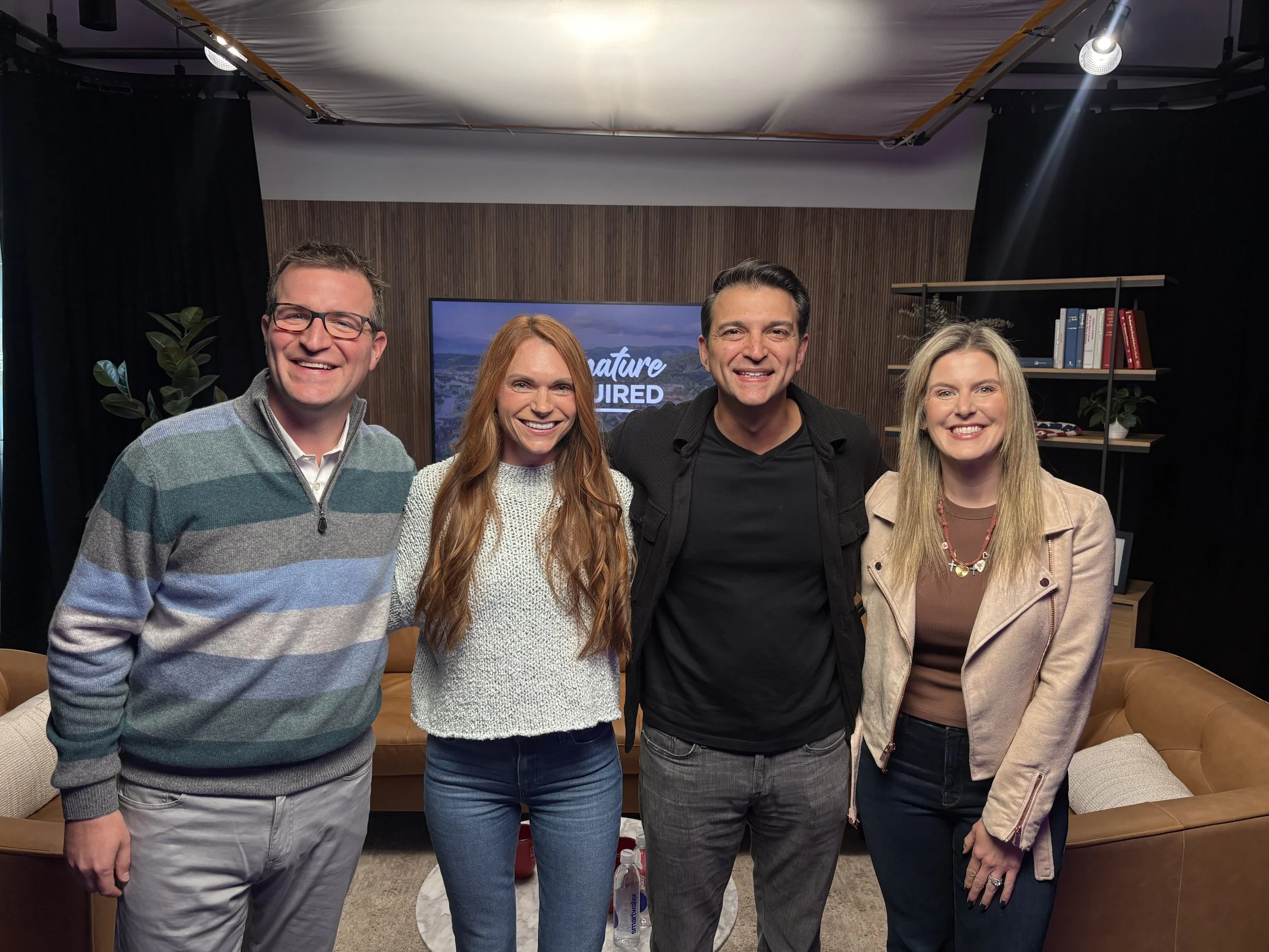 Four smiling people standing closely together in a room with a TV screen in the background that has text 'Signature Required' on it, and bookshelves on the right side.