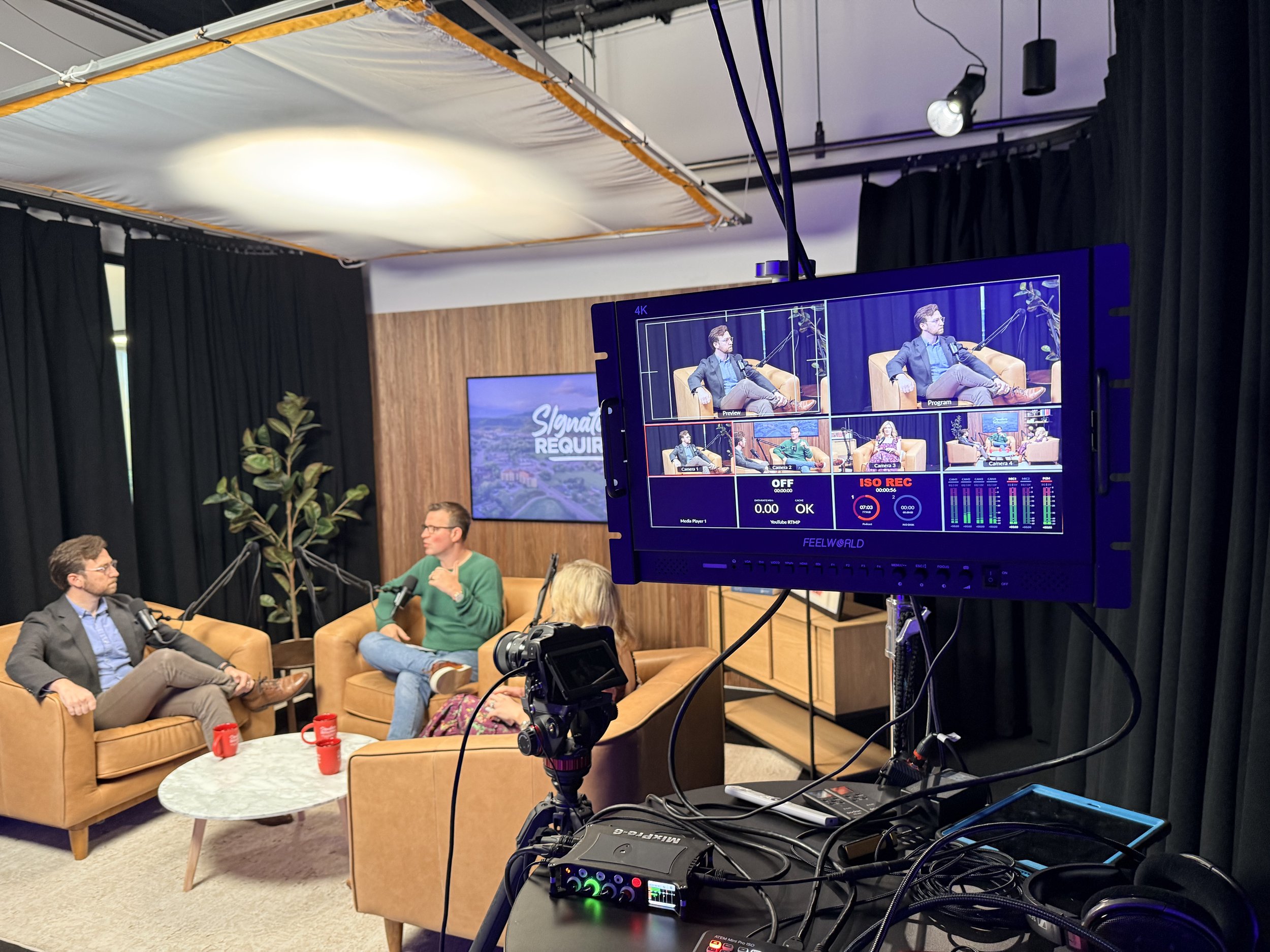 Recording studio or podcast setup with two men seated on tan couches engaged in conversation, microphone setup, a large monitor displaying video feed, and technical equipment in the foreground.