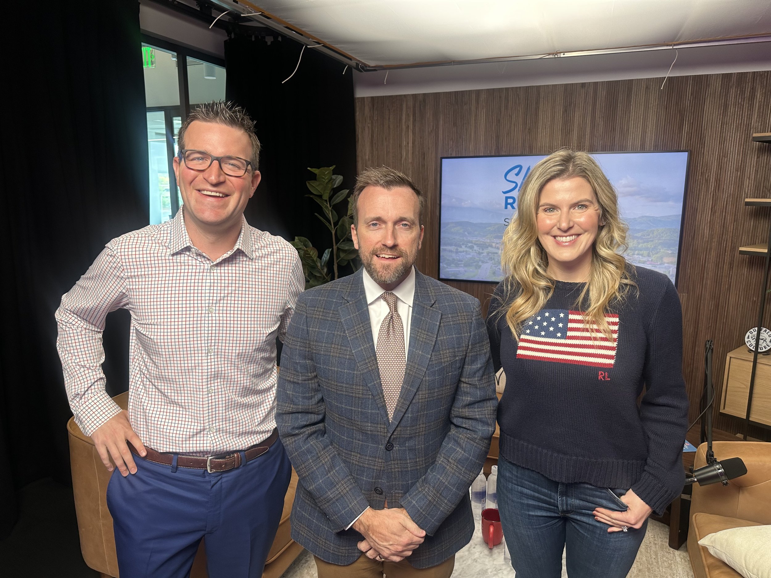 Three people standing side by side indoors, smiling at the camera. The person on the left is a man with glasses wearing a checked shirt, the person in the middle is a man with a beard wearing a checkered blazer, and the person on the right is a woman