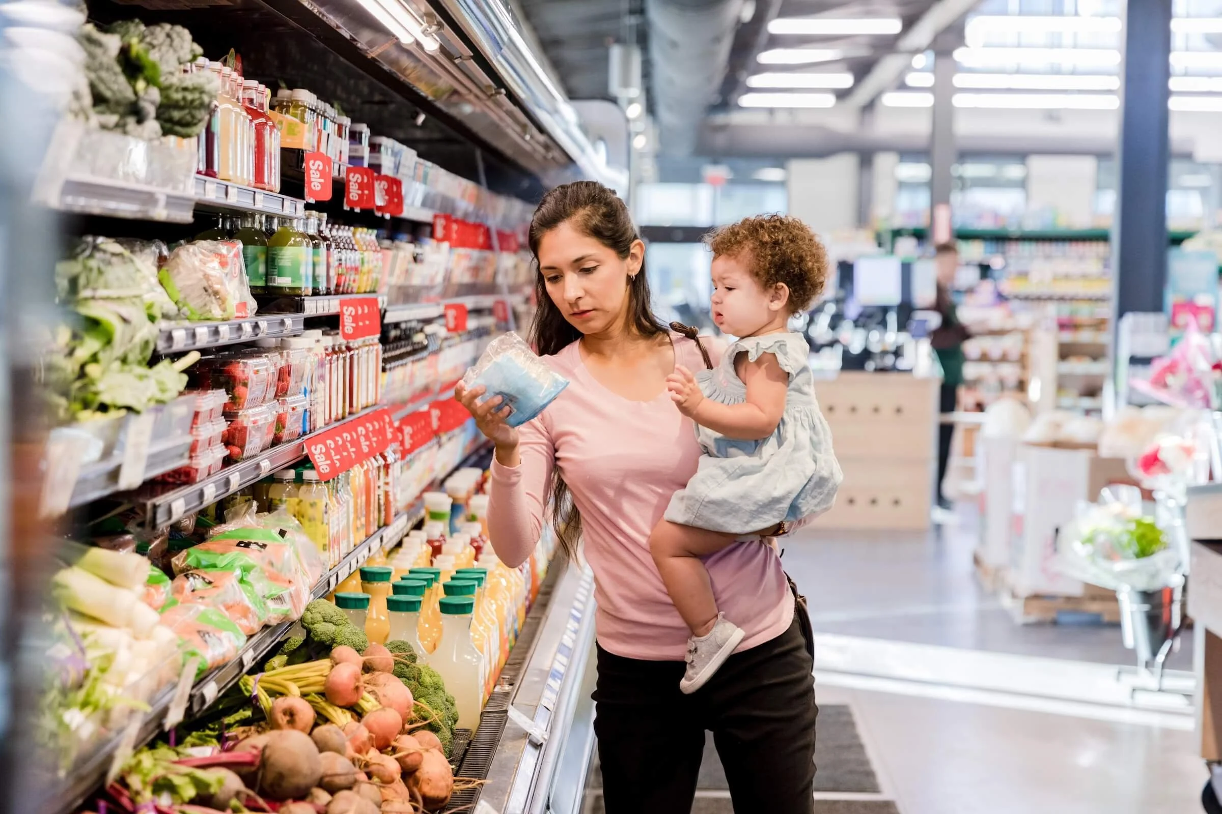 A woman holding a child shops at a grocery store, examining a packaged product in the produce aisle with various fruits, vegetables, and juice bottles on the shelves.