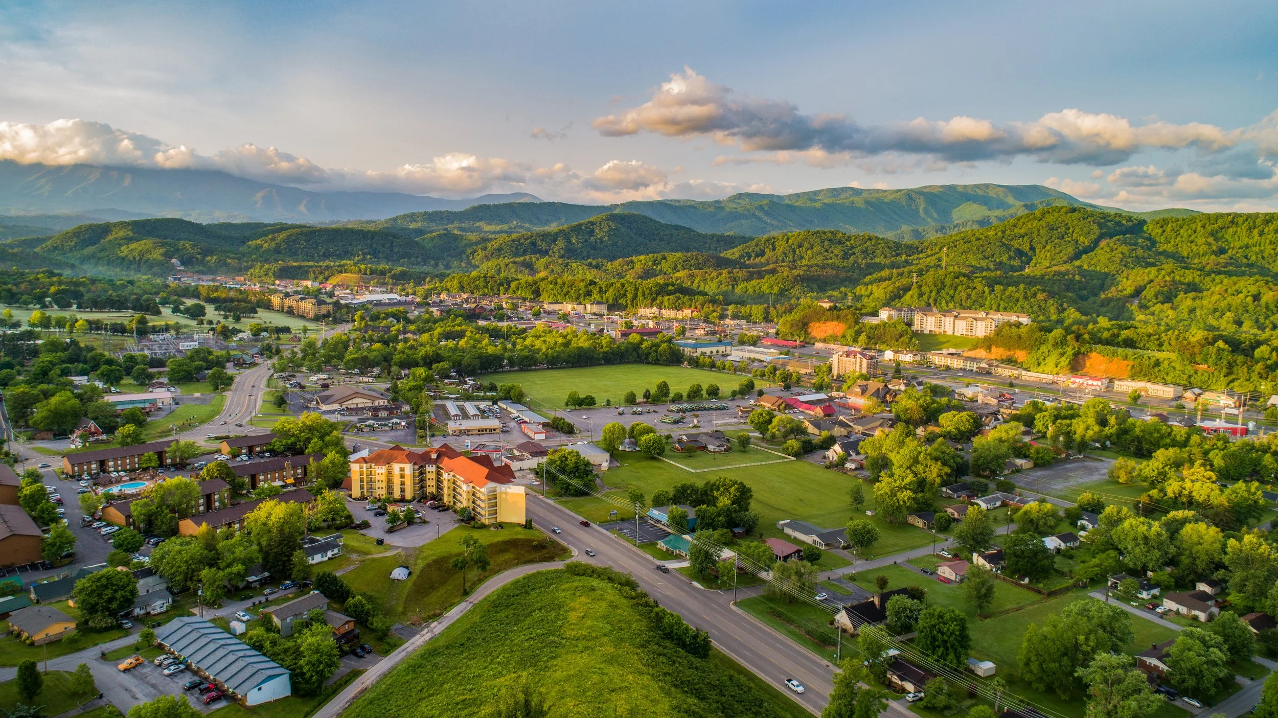 Aerial view of a town surrounded by green hills and mountains under a partly cloudy sky.