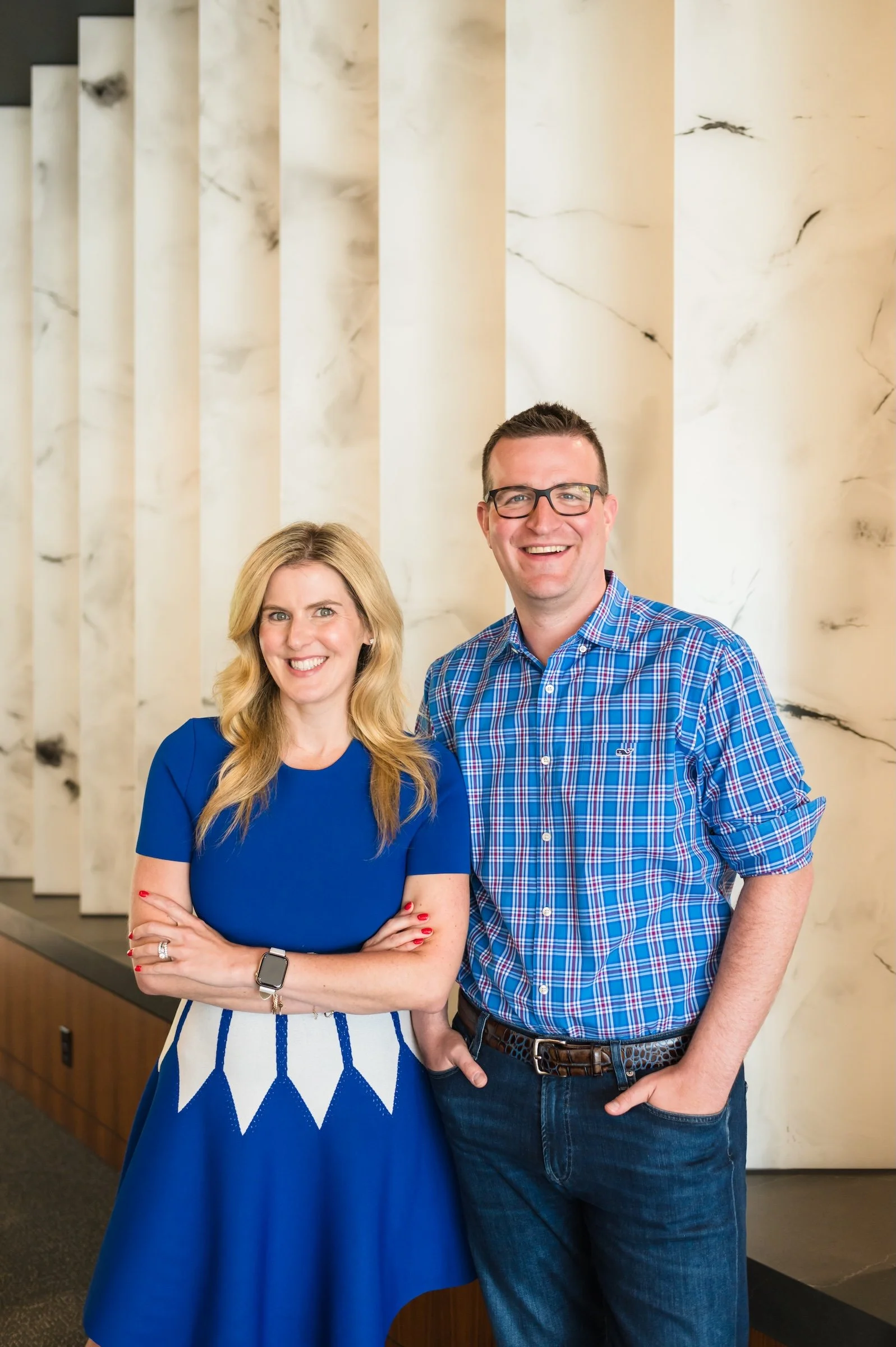 Two people smiling, standing in front of a cream marble wall. The woman is wearing a blue dress, and the man is in a blue checked shirt and jeans.