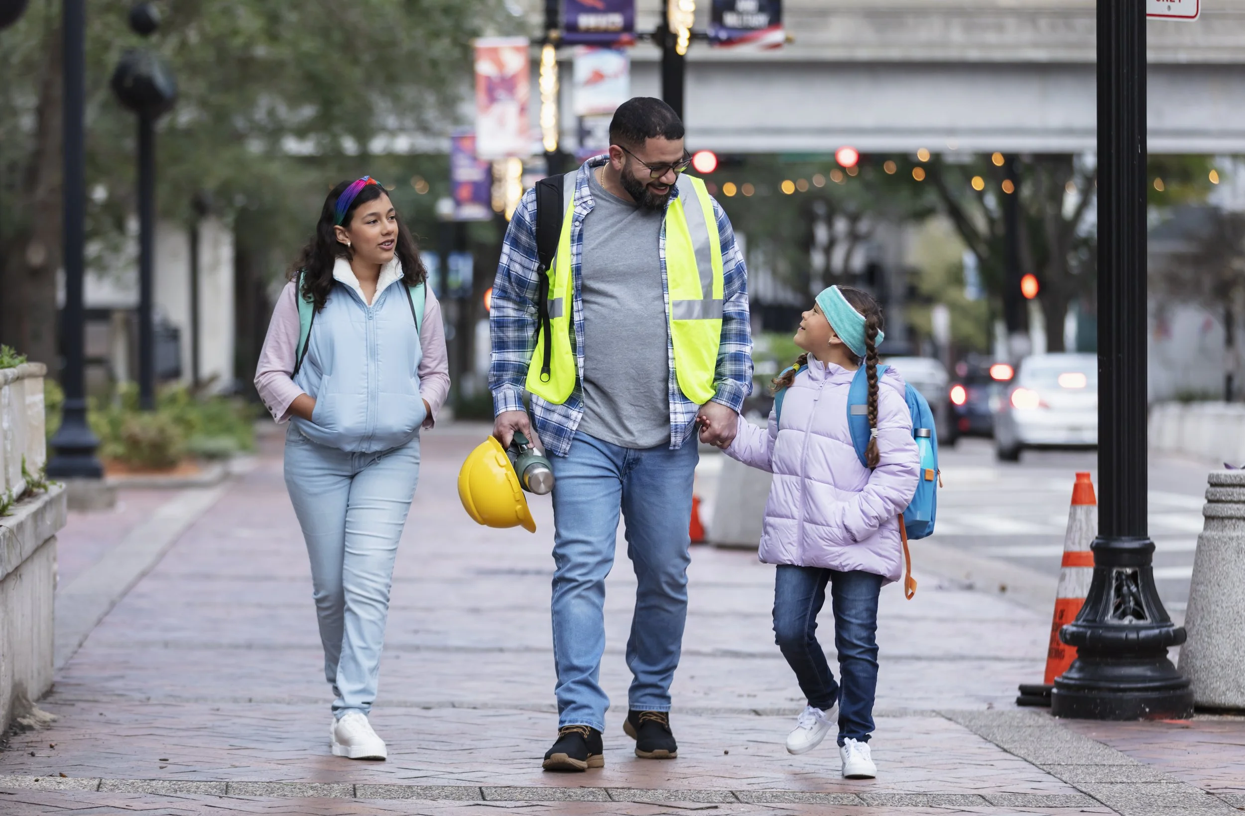 A man wearing a safety vest and carrying a yellow hard hat walks with two young girls on a city sidewalk.