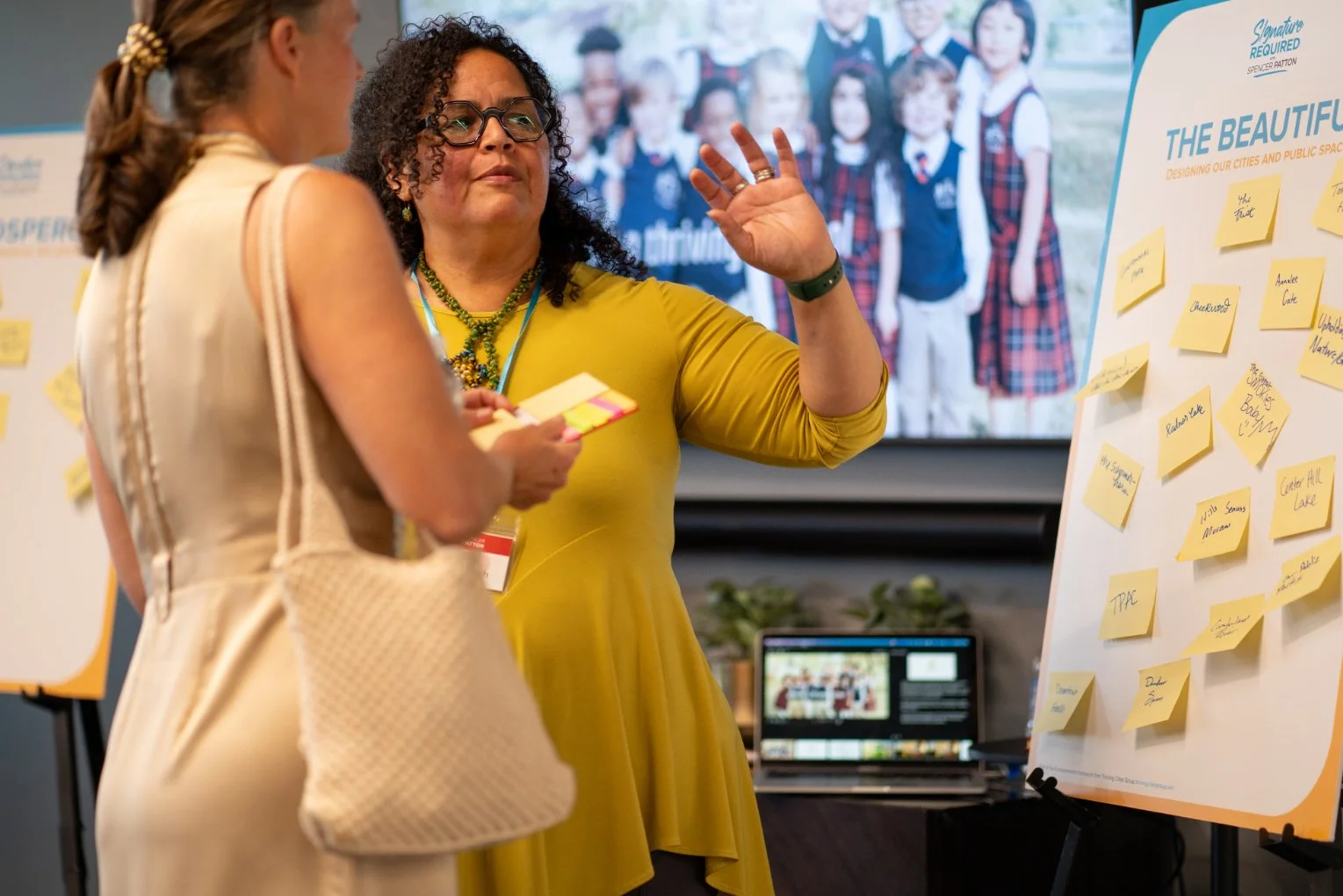 Two women engaged in a discussion at a conference, with a large brainstorming board covered in yellow sticky notes and a group of children on a screen in the background.