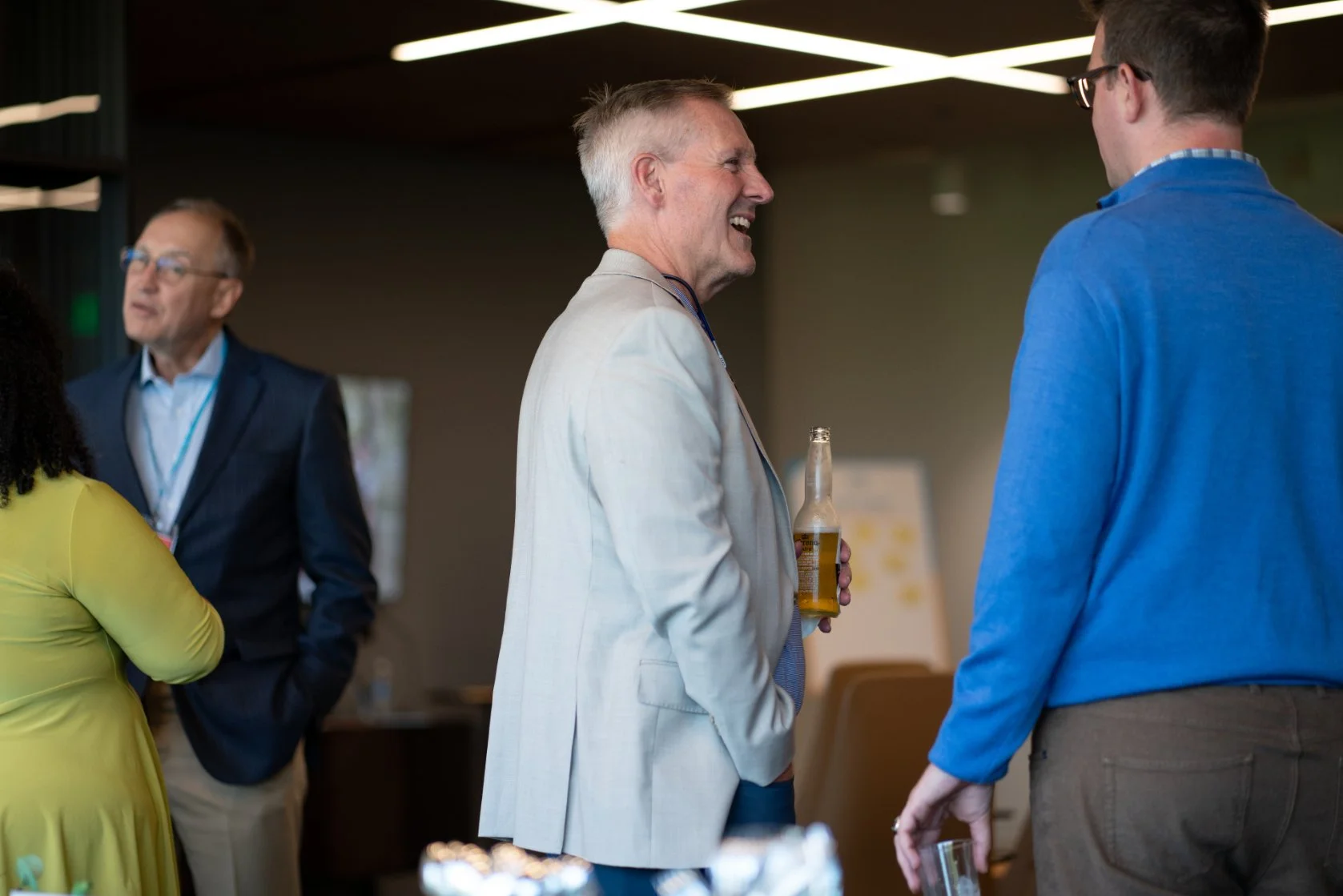 People talking at a social gathering, including a man in a white suit holding a beer, smiling and conversing with a man in a blue shirt, with others in the background.