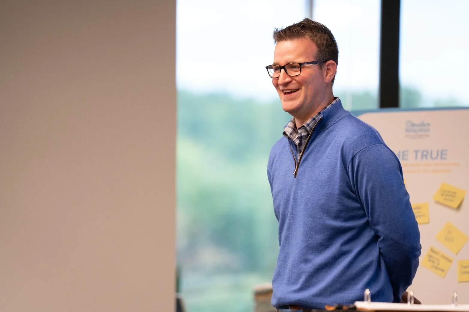 A man in glasses and a blue quarter-zip sweater smiling during a presentation or meeting, with a banner featuring post-it notes behind him.