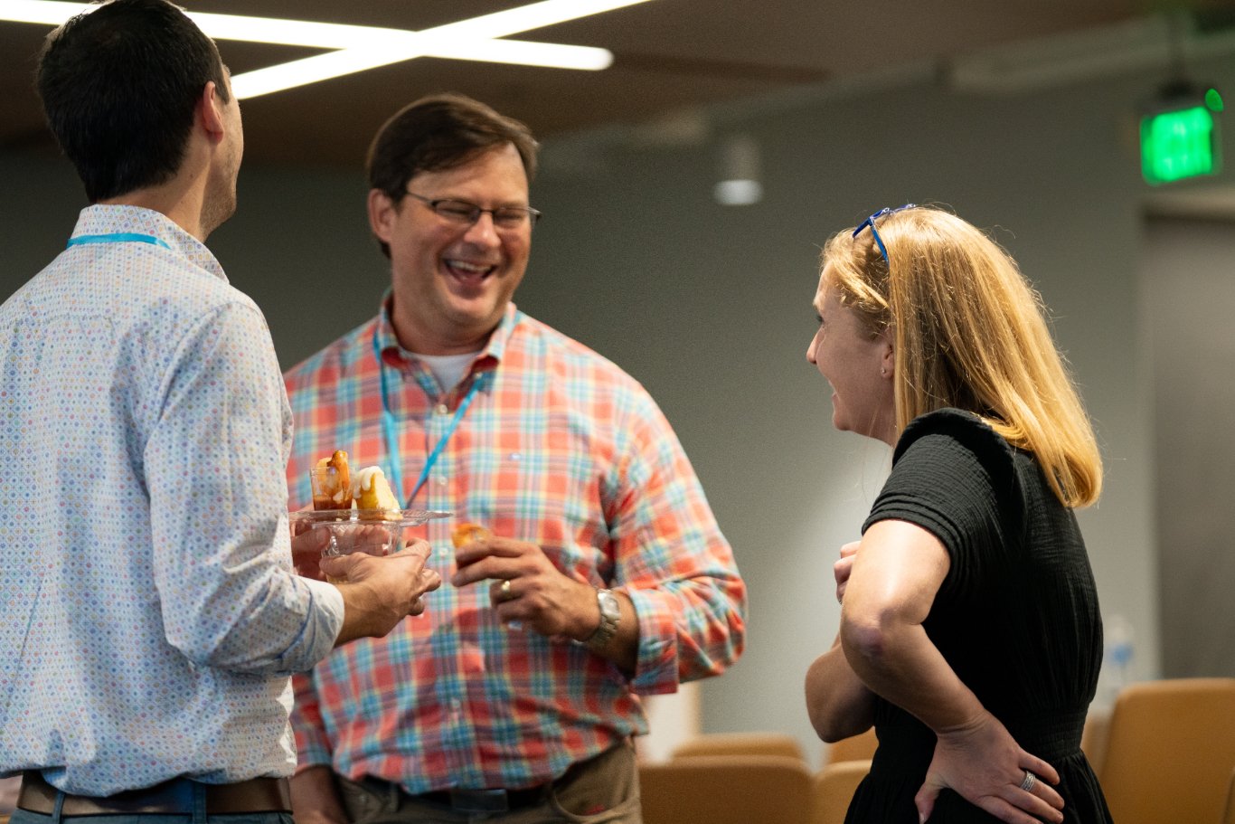 Three people laughing and talking at an indoor event, one person in the middle holding a plate of food, all casually dressed.