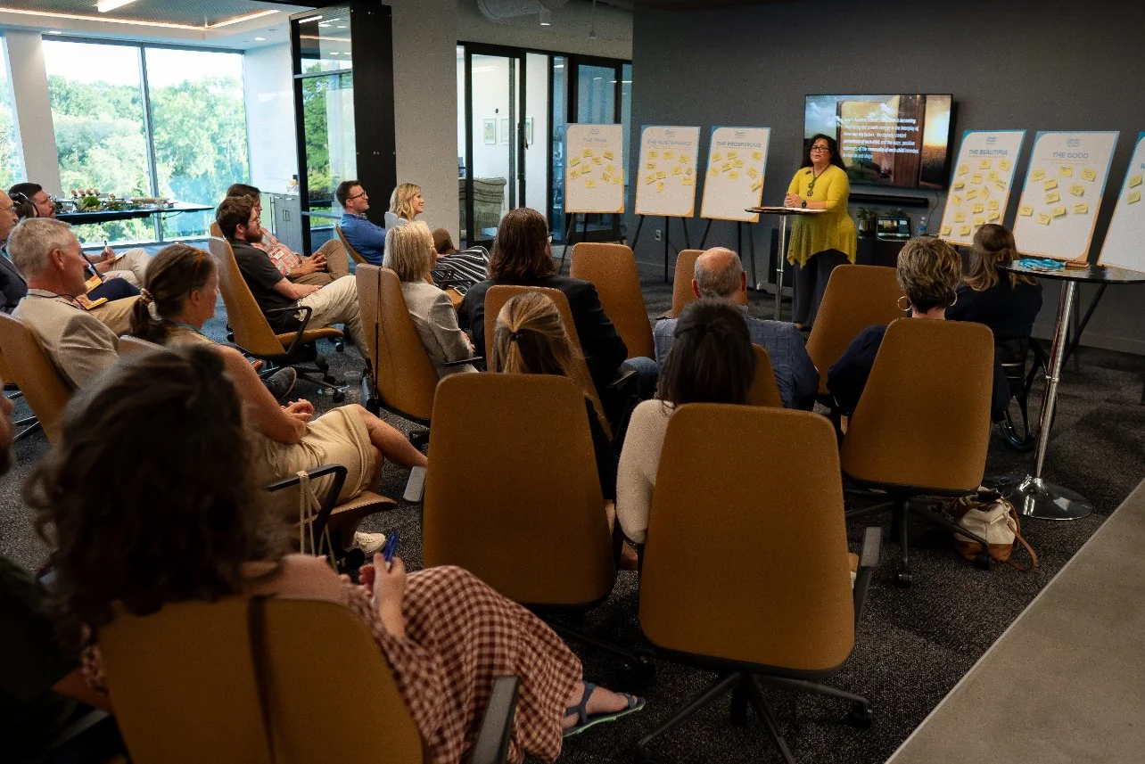 A woman in a yellow dress presenting to a seated audience in a conference room, with whiteboards filled with yellow sticky notes and a screen displaying a slide, during a workshop or seminar.