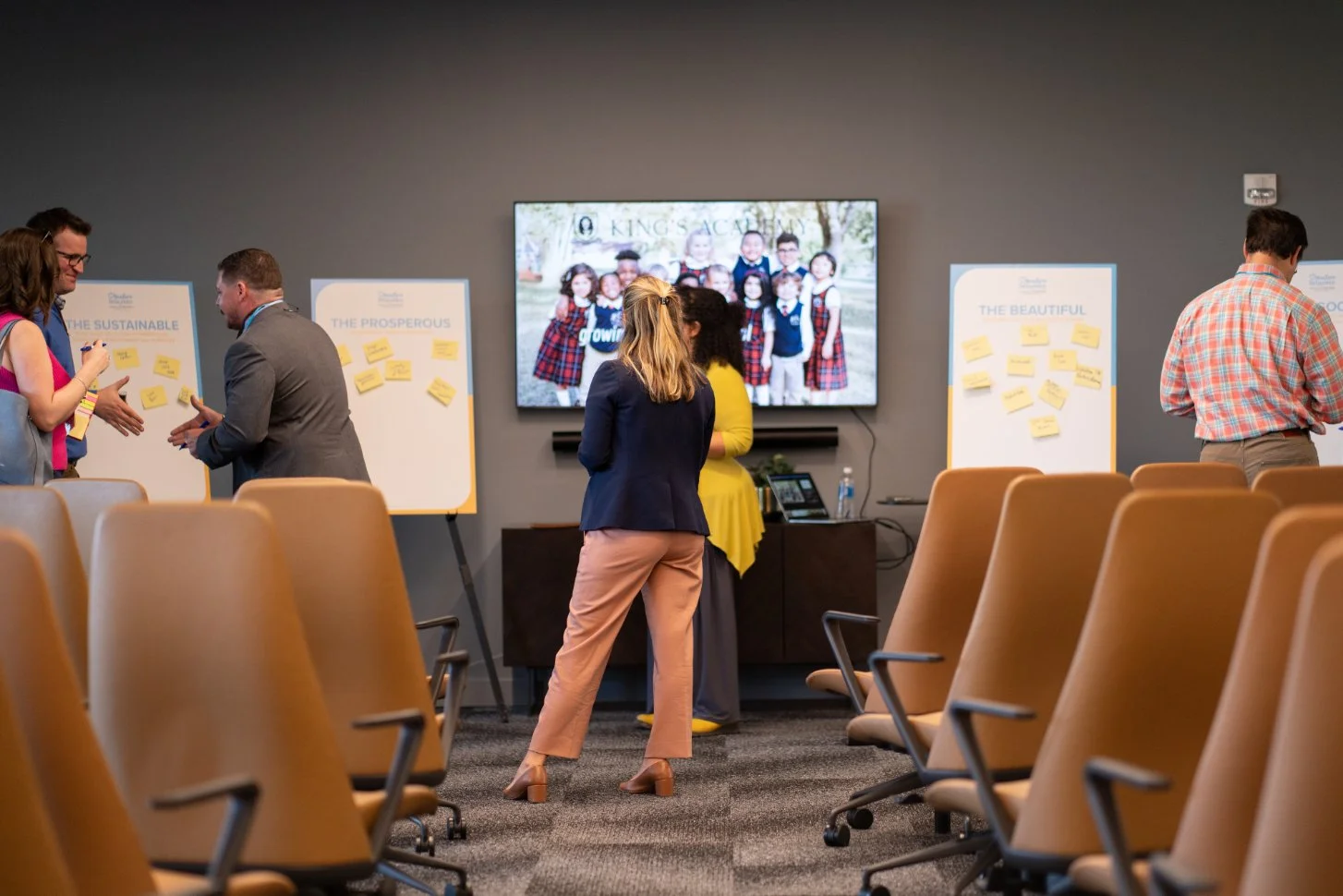 People in a conference room engaging in discussions near whiteboards with sticky notes, with a large group photo on a TV screen in the background.