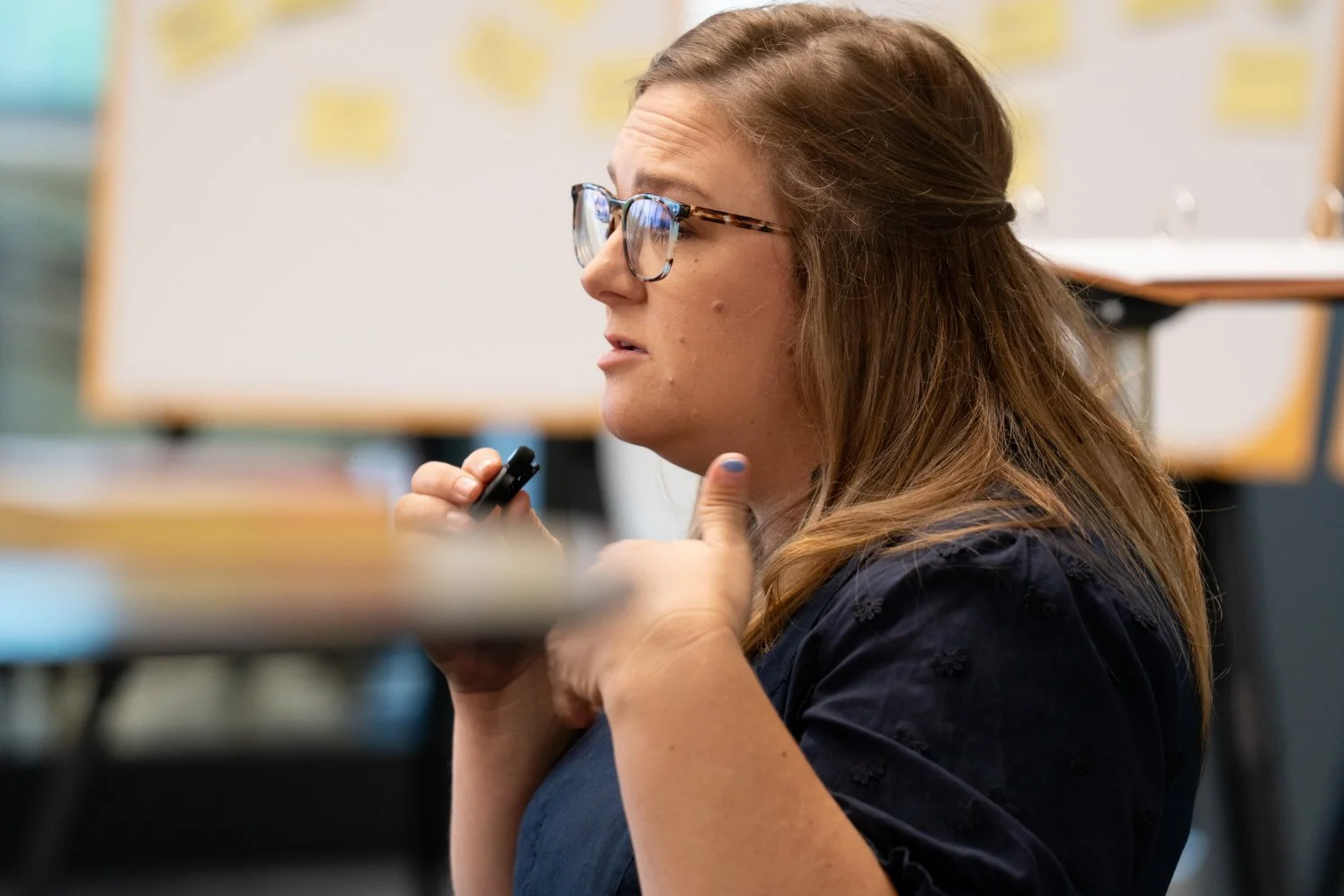 A woman with glasses and light brown hair, holding a marker near her neck, in a classroom with a whiteboard in the background.
