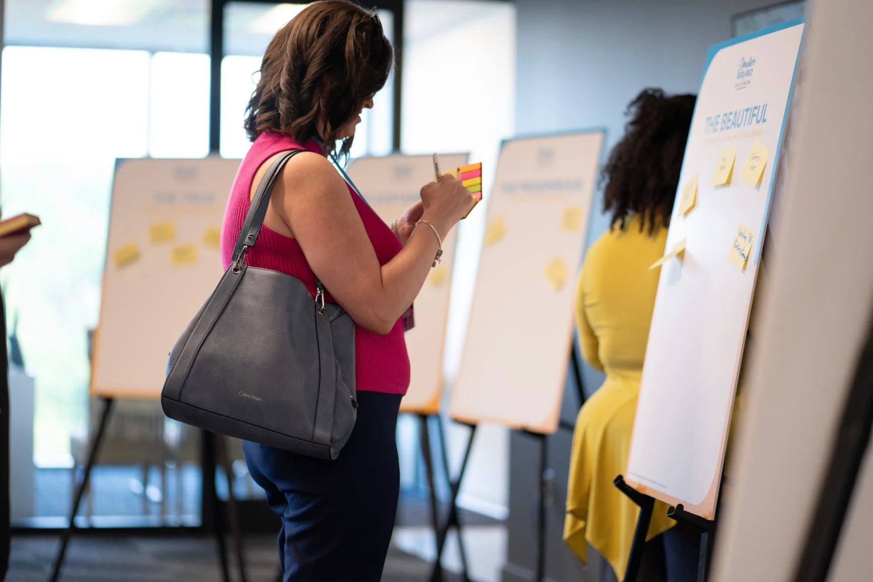 A woman in a sleeveless pink top and dark pants is looking at a sticky note on a whiteboard at a conference or workshop. She has shoulder-length brown hair, a grey handbag on her shoulder, and is taking notes. There are multiple whiteboards with stic
