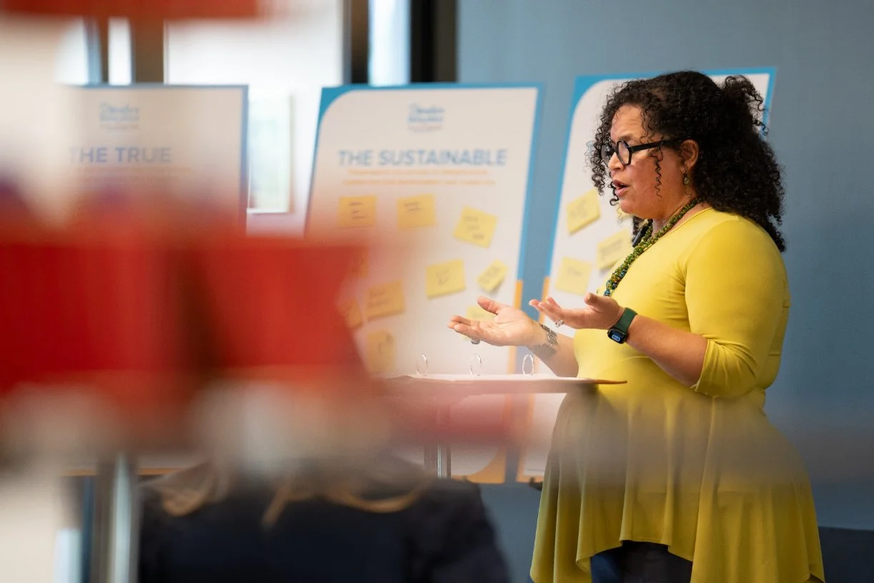 A woman with curly hair and glasses is speaking in front of presentation boards with sticky notes, wearing a yellow dress and a beaded necklace.