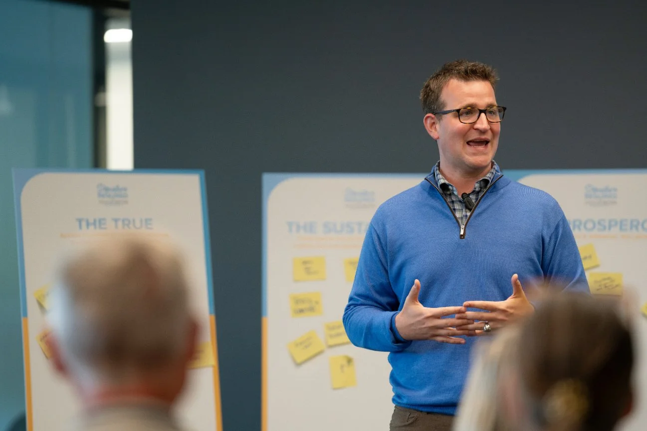 Man wearing glasses and a blue sweater giving a presentation in front of boards with yellow sticky notes, with an audience in the foreground.