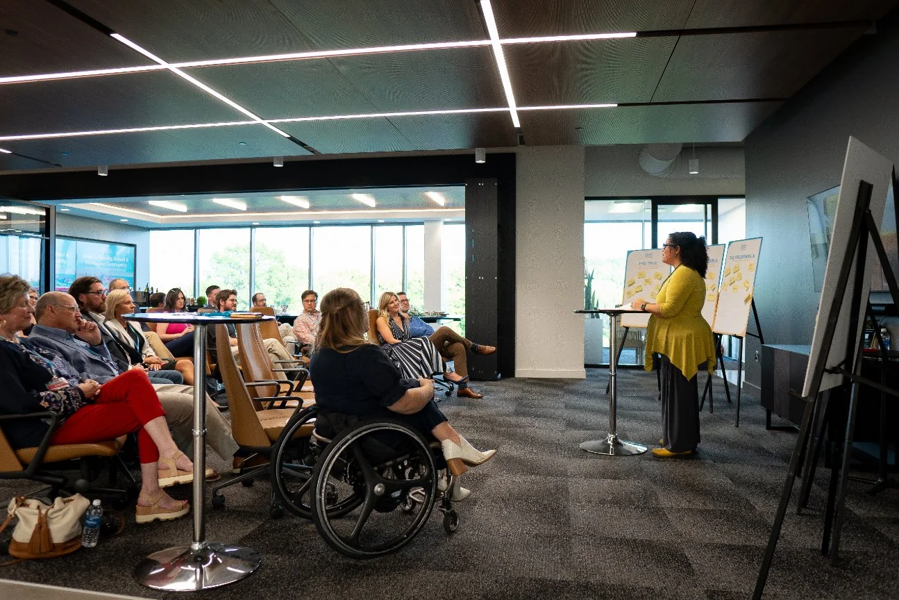 A woman in a yellow coat gives a presentation to an audience in a modern conference room with large windows and ceiling lights.
