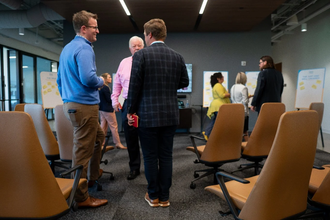 People gathered and conversing in a modern conference room with brown leather chairs and whiteboards.