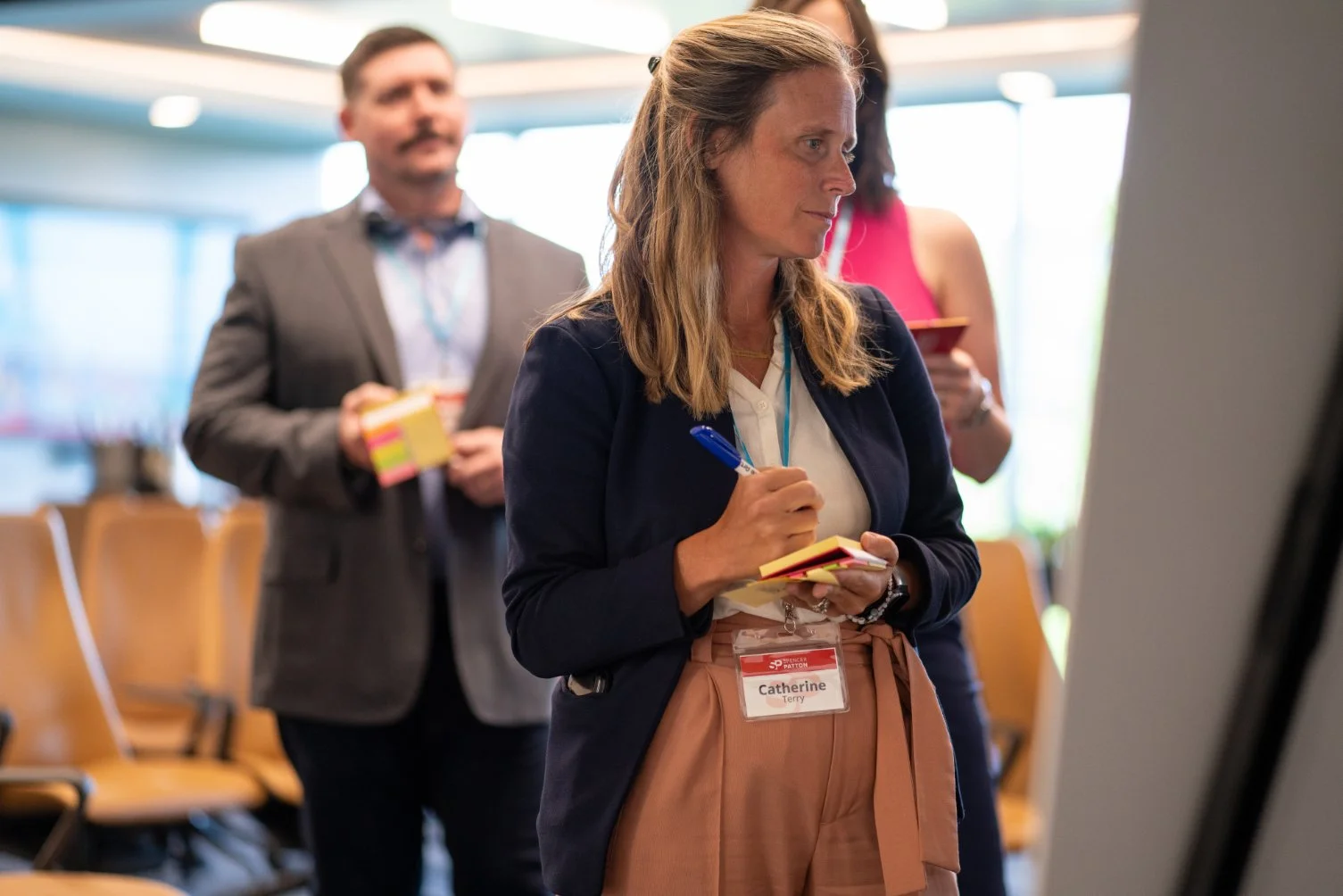 Woman wearing a name tag, taking notes during a conference or meeting, with people in the background holding papers and standing near empty chairs.