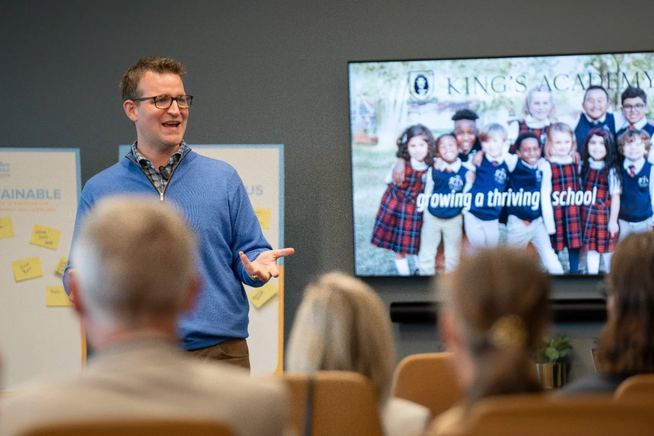 A man giving a presentation in a classroom or conference room, with a large screen showing students in school uniforms and the words "Gowing a thriving school." The man is wearing glasses and a blue sweater, and is gesturing with his right hand. Seve