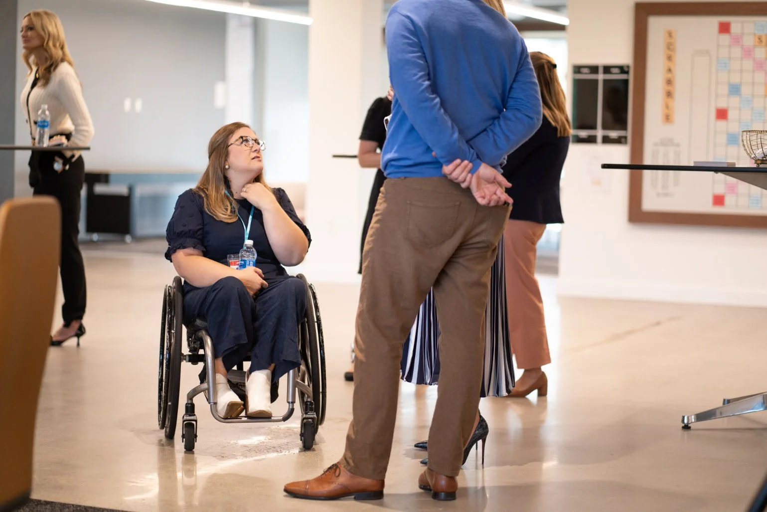 Group of people having a conversation in an indoor setting, with a woman in a wheelchair holding a water bottle and listening to a man in a blue blazer speaking, all casually dressed.