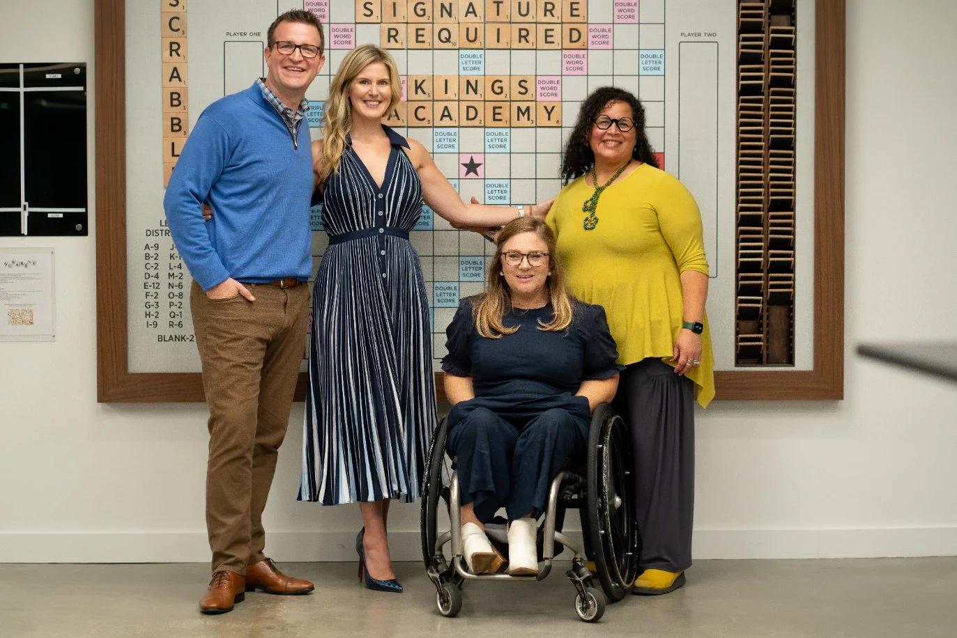 Four people, including one woman in a wheelchair, standing in front of a Scrabble board.