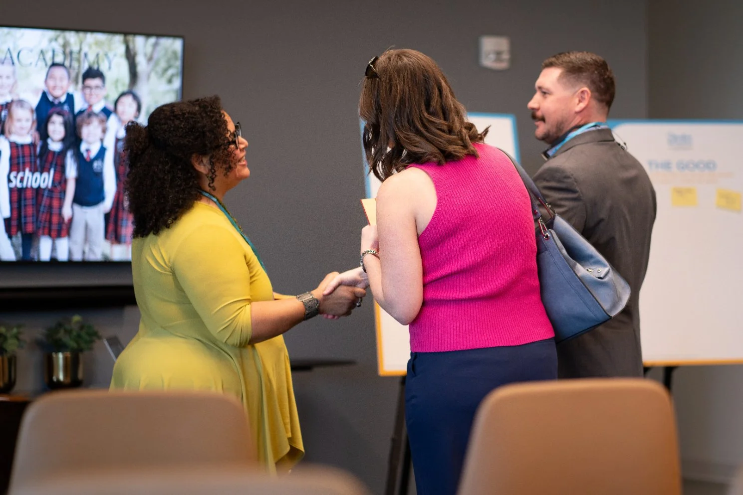 Three adults engaged in conversation at a professional event or conference, with a large screen displaying children in school uniforms in the background.