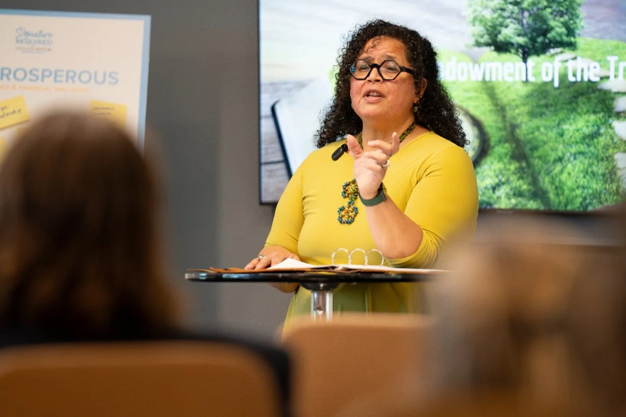 A woman with curly dark hair, glasses, and a yellow top speaking at a presentation, with an audience in the foreground and a large screen behind her displaying greenery and text.