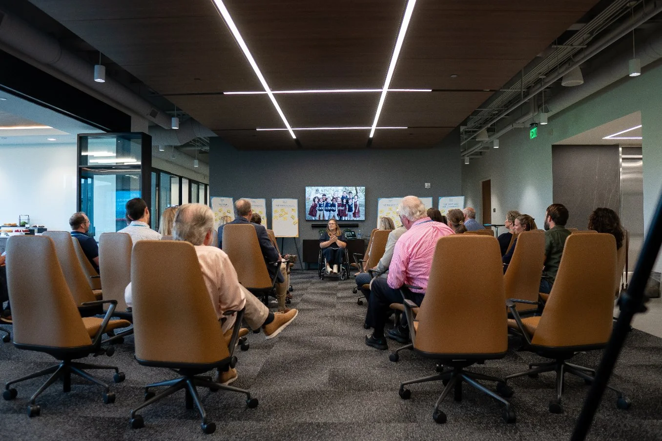 People seated in a conference room listening to a speaker at the front, with a large screen displaying a presentation.