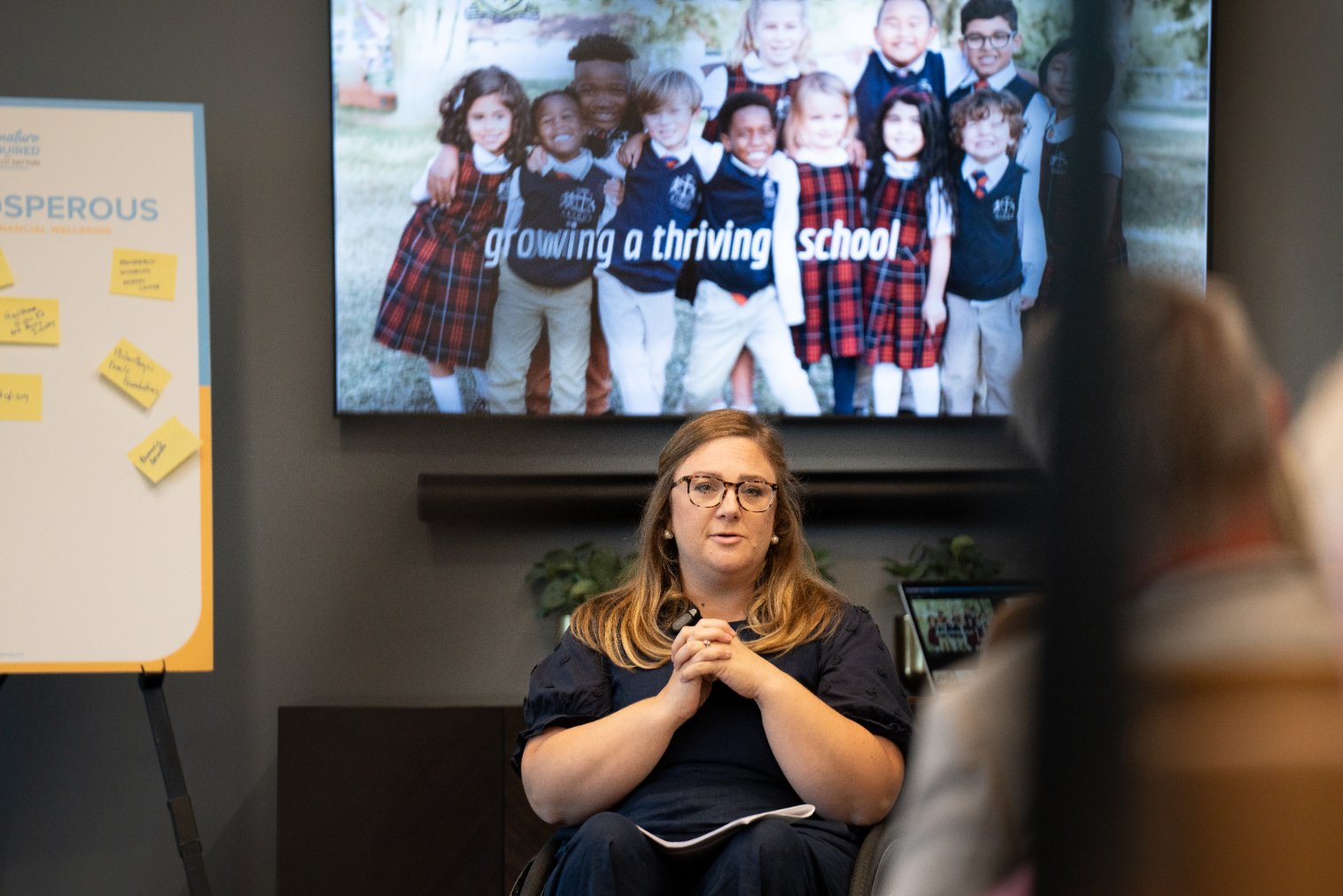 A woman with glasses and long hair speaking in a meeting room with a large screen behind her showing a photo of diverse children in school uniforms and the words "growing a thriving school." An audience member is visible in the foreground.
