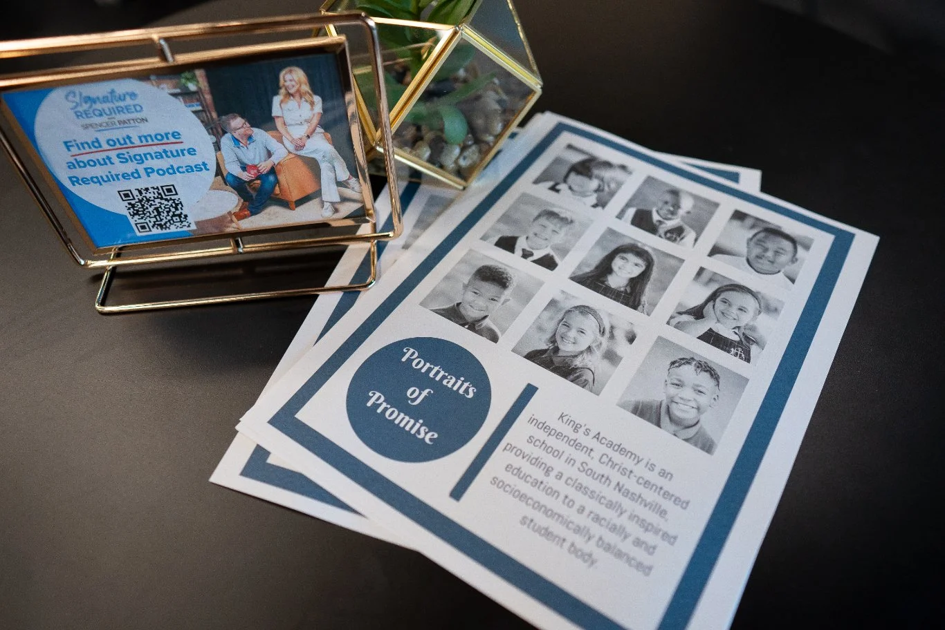 Brochures titled 'Portraits of Promise' featuring photos and information about King's Academy, an independent, Christ-centered school with a diverse student body, placed on a table beside a display with a photo of two students and a QR code for the '