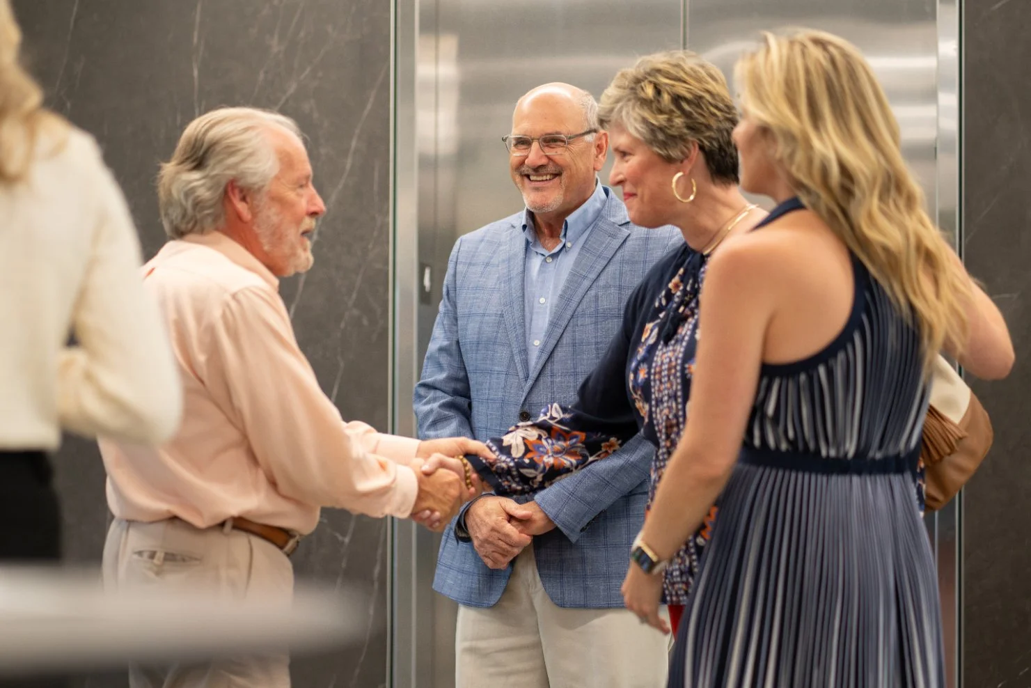A group of well-dressed people greeting each other in an elevator, including a man and woman shaking hands and smiling.