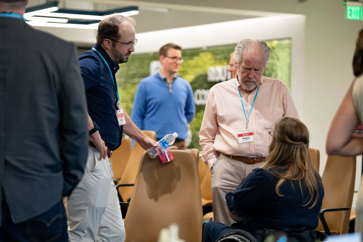 A group of people engaged in conversation in a modern, well-lit conference room, with chairs and an indoor greenery background.