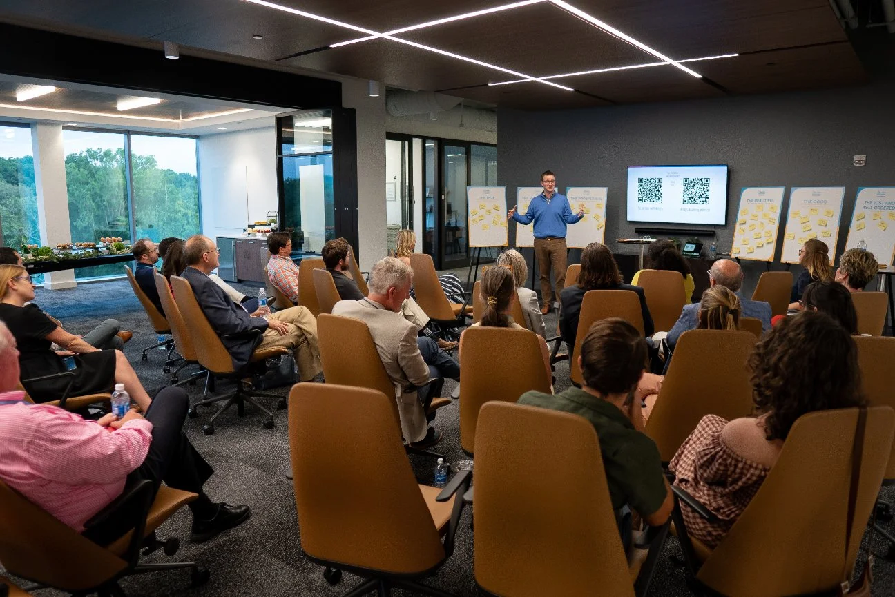 A man in a blue shirt giving a presentation to a group of people in a modern conference room, with large windows showing a green outdoor view, and screens displaying QR codes behind him.
