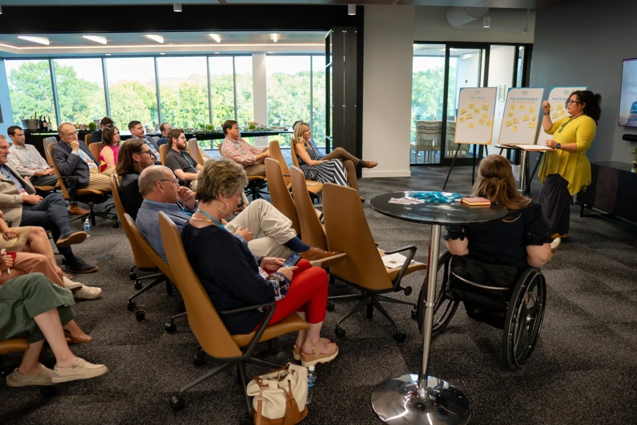 A woman in a yellow dress presents in a conference room filled with seated attendees. The room has large windows overlooking trees, and there are presentation boards with sticky notes and a screen on the wall.