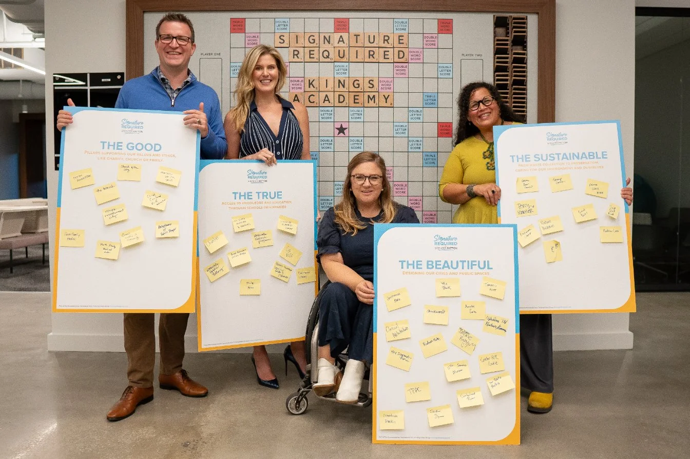 Group of five women and men holding posters with values written on sticky notes, standing in front of a crossword puzzle board labeled 'SIGNATURE REQUIRED'.