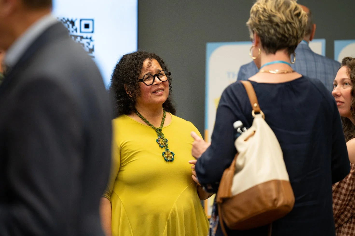 A woman in a yellow dress with glasses and a green necklace talking to a woman with short black hair, gold earrings, and a dark blazer in a professional setting.