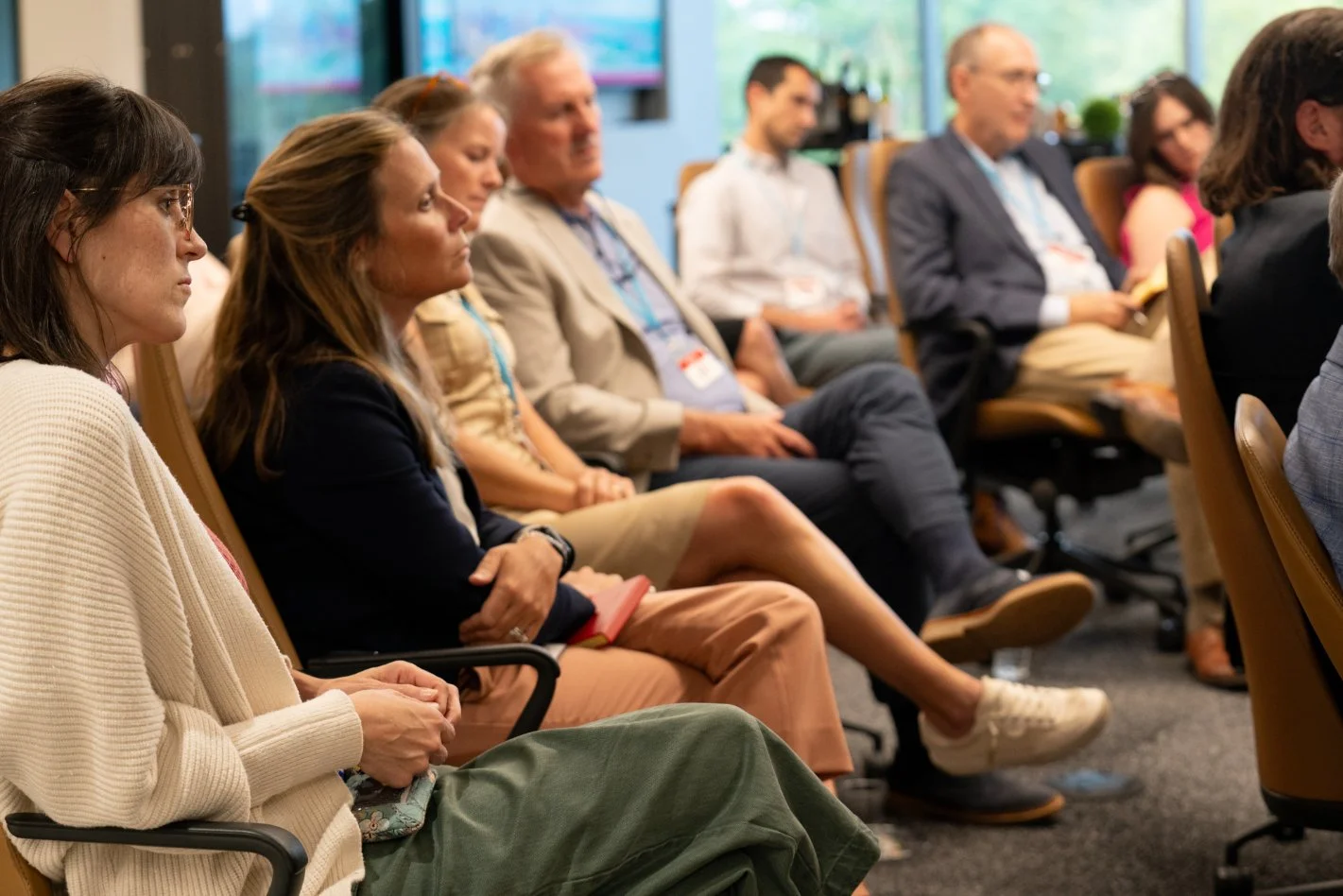 People attending a conference or seminar, sitting in chairs, listening attentively in a well-lit room with large windows.