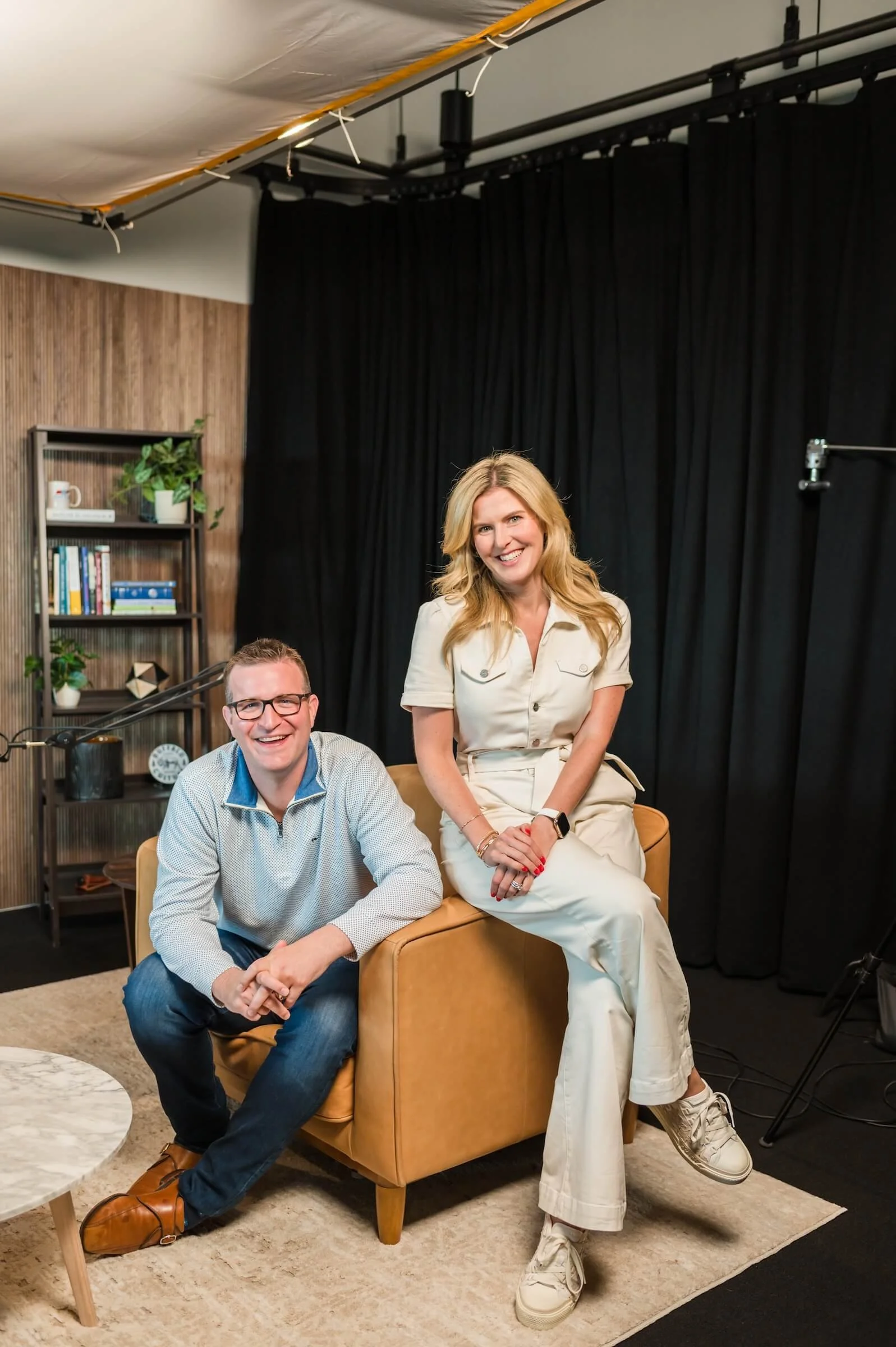 A man and woman smiling and sitting on a tan armchair in a room with dark curtains and wooden wall. The man is sitting on the armchair's arm, wearing glasses and a light-colored shirt. The woman is sitting on top of the chair, wearing a cream-colored