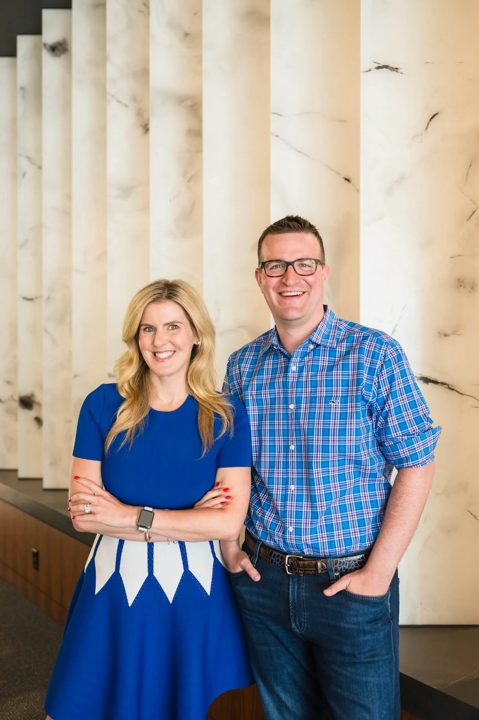 A woman and a man smiling, standing indoors in front of a marble wall with vertical columns. The woman has blonde hair, is wearing a blue dress, and the man has short dark hair and glasses, is wearing a blue plaid shirt and jeans.
