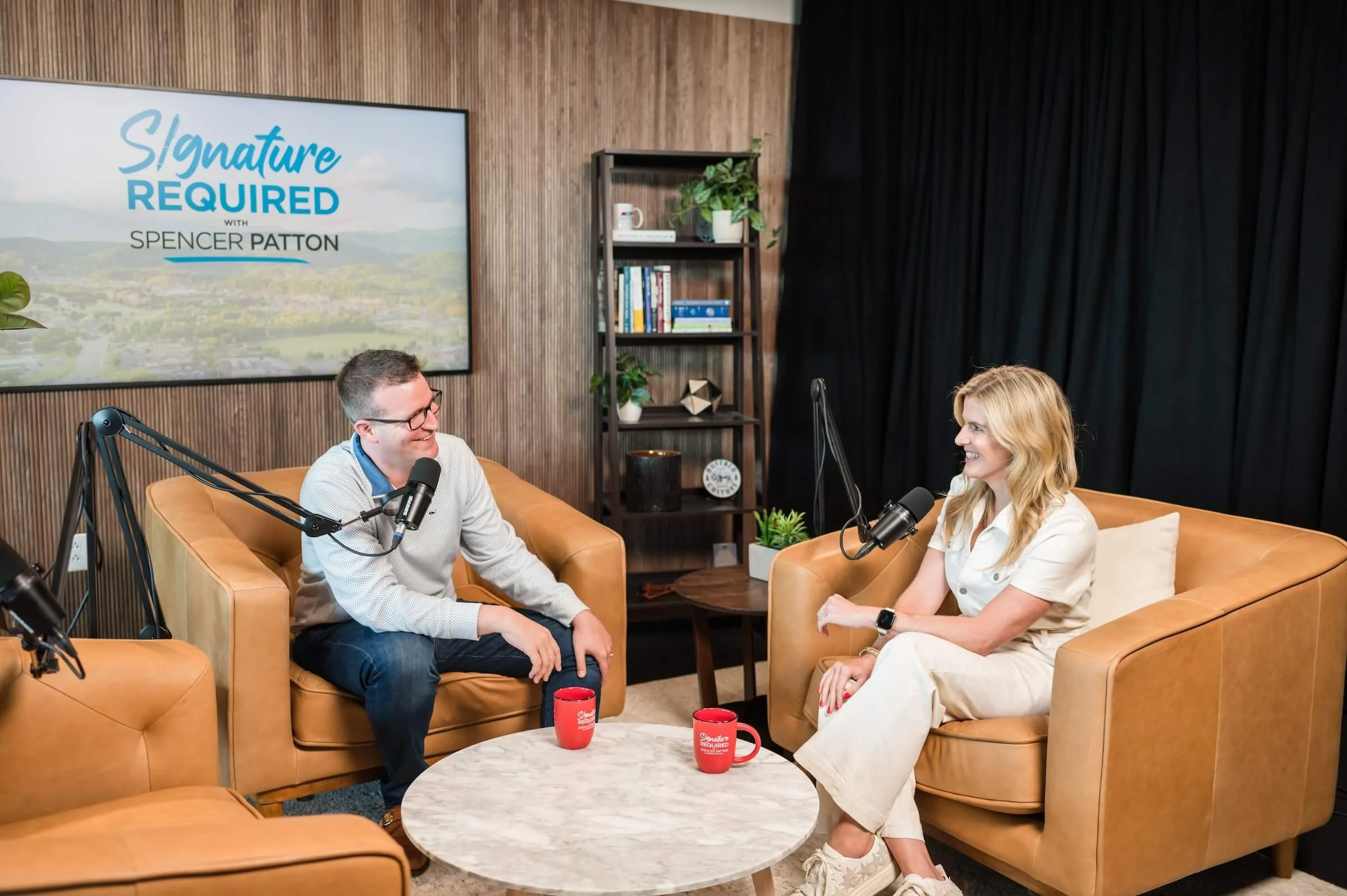 Two people sitting on tan leather couches in a podcast studio, engaging in conversation with microphones, with a screen behind them displaying 'Signature REQUIRED with Spencer Patton', a bookshelf with decor, and black curtains.