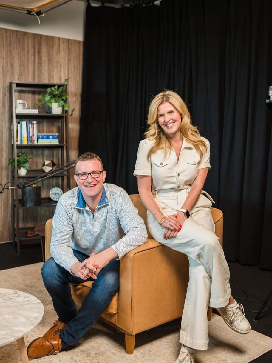 A man and woman smiling and sitting in a modern, cozy room with black curtains and wooden accents.