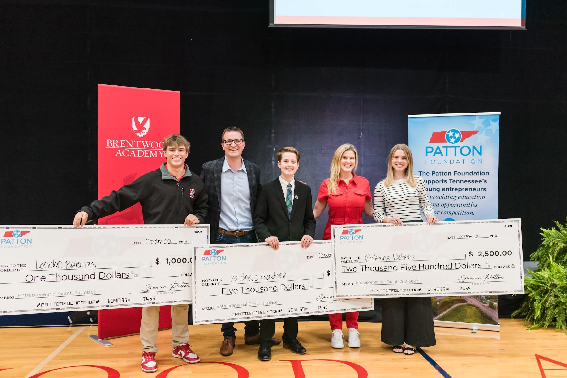Group of six people on stage holding oversized checks at an award ceremony, with banners for Brentwood Academy and the Patton Foundation in the background.