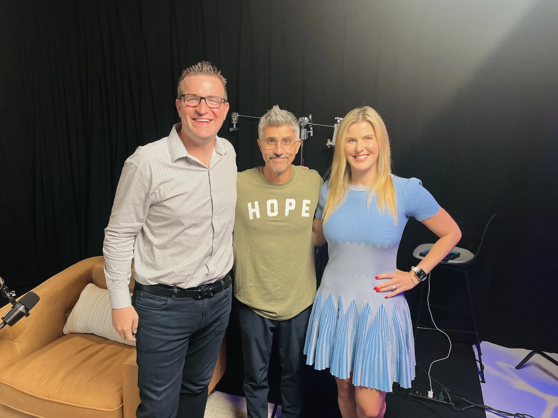 Three smiling people standing close together in a studio, with black curtains behind them and a mustard-colored couch on the left. The man on the left is wearing glasses, a white patterned shirt, and dark pants. The man in the middle is wearing a green T-shirt with 'HOPE' written on it, and glasses. The woman on the right has long blonde hair, is wearing a light blue dress, and has her hand on her hip.