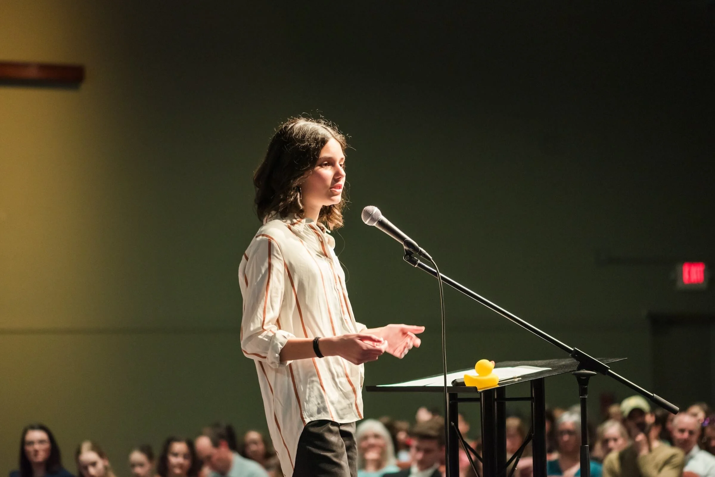 A woman stands at a podium with a microphone, speaking in front of an audience in a dimly lit room.