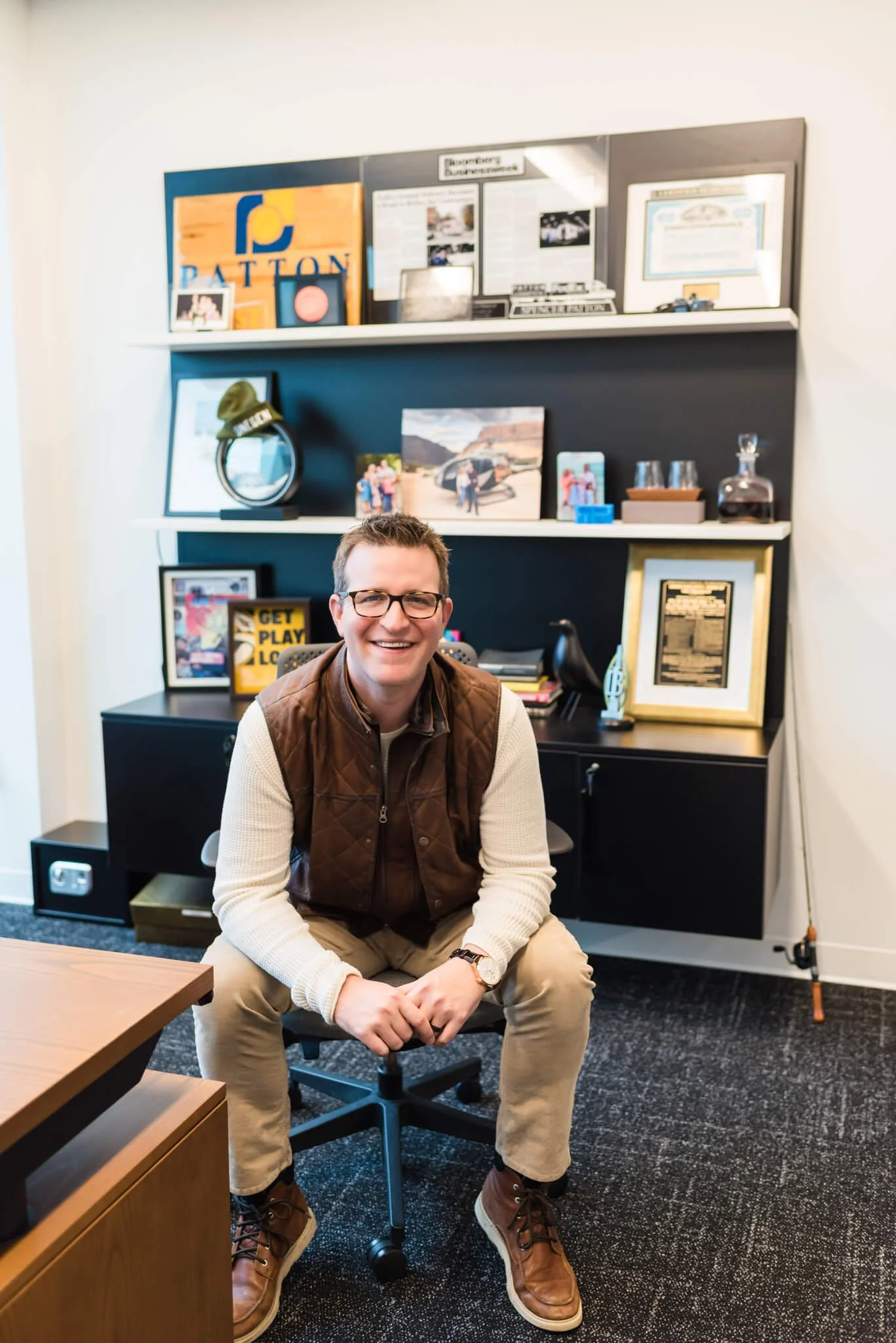 A man with glasses, wearing a brown vest and beige pants, smiling and sitting on an office chair in a modern office space with a black and white wall-mounted bookshelf behind him decorated with framed photos, awards, and decorative items.