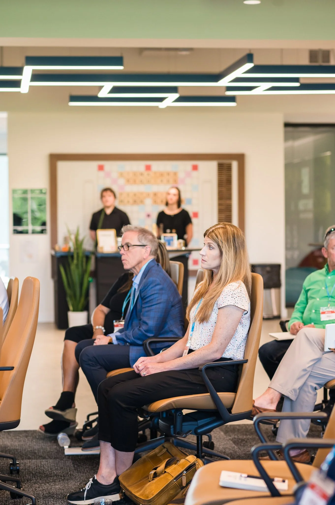 A group of people are sitting in conference chairs attending a presentation or seminar in a modern conference room, with two women standing at a table in the background.